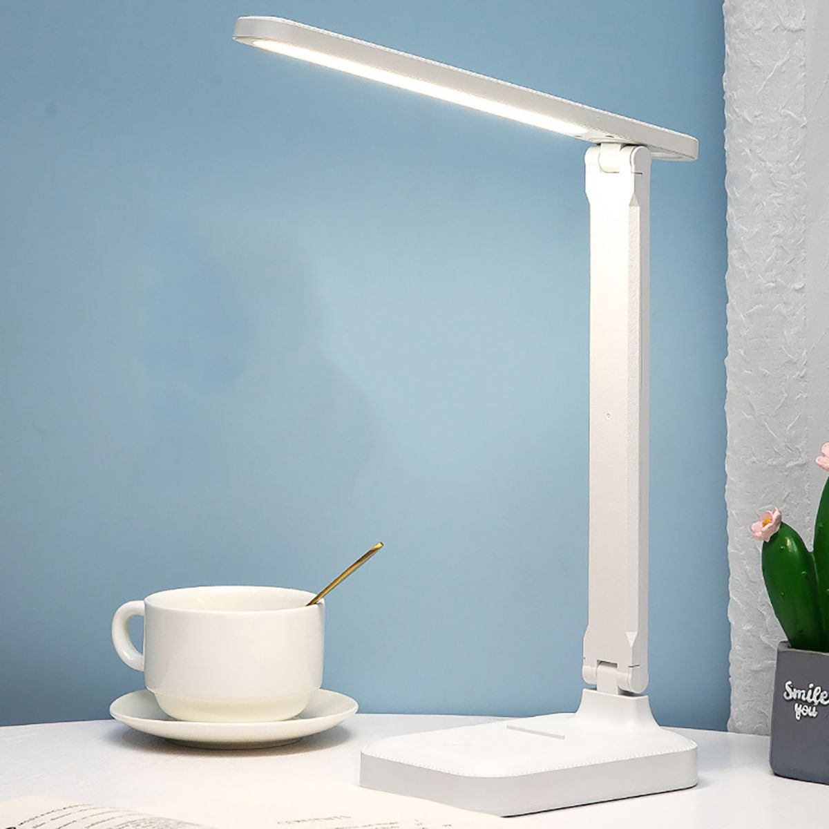 Weiße Schreibtischlampe mit flexiblem Kopf. Weißer Sockel. Blauer Hintergrund. Tasse und Kaktus.