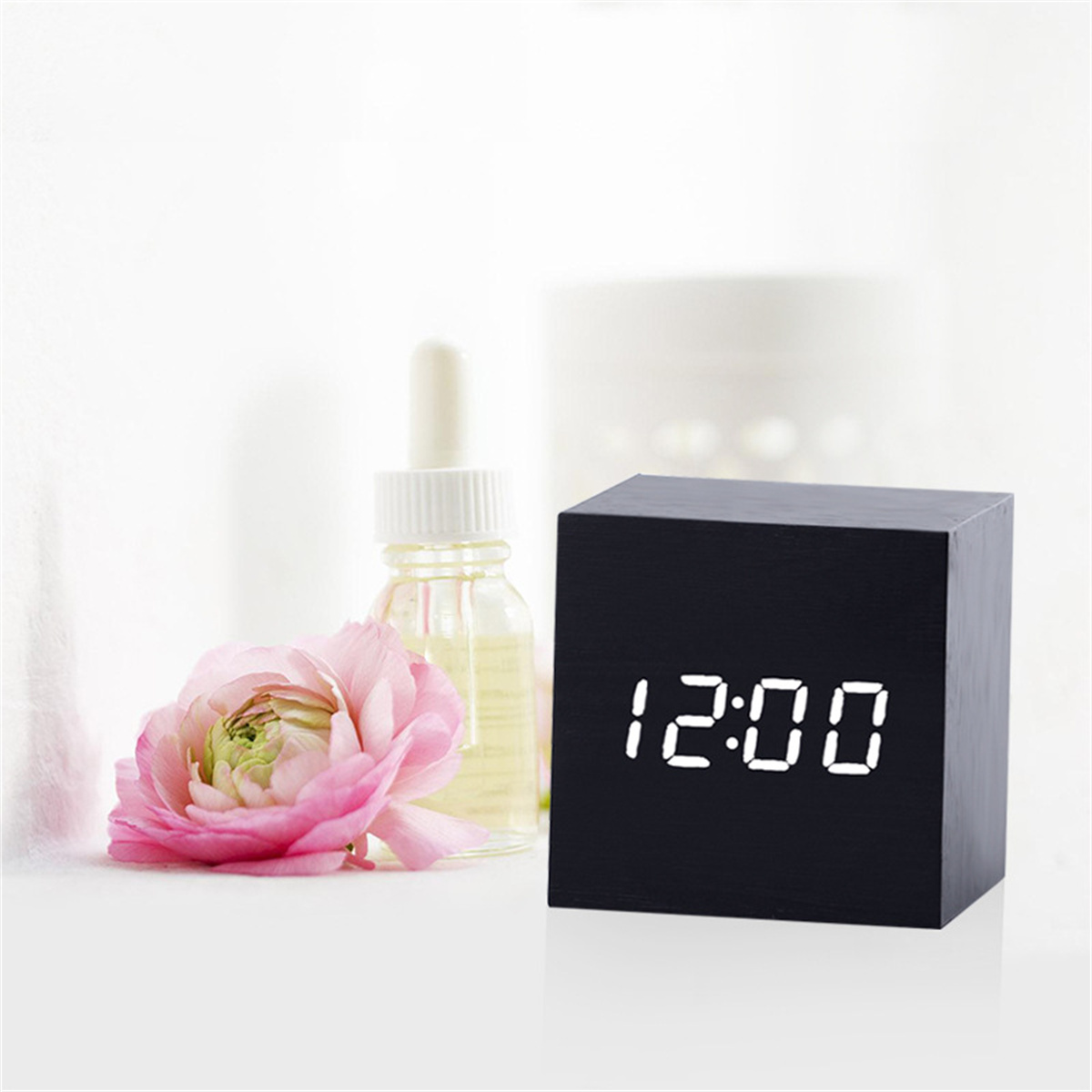 Schwarze Uhr mit weißen Zahlen, die 12:00 anzeigt, neben einer rosa Blume und einer Ölflasche.
