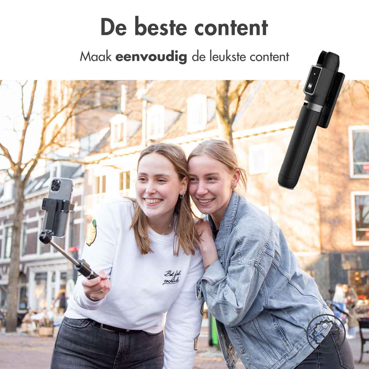 Two women smiling, taking a selfie using a selfie stick in a city street.