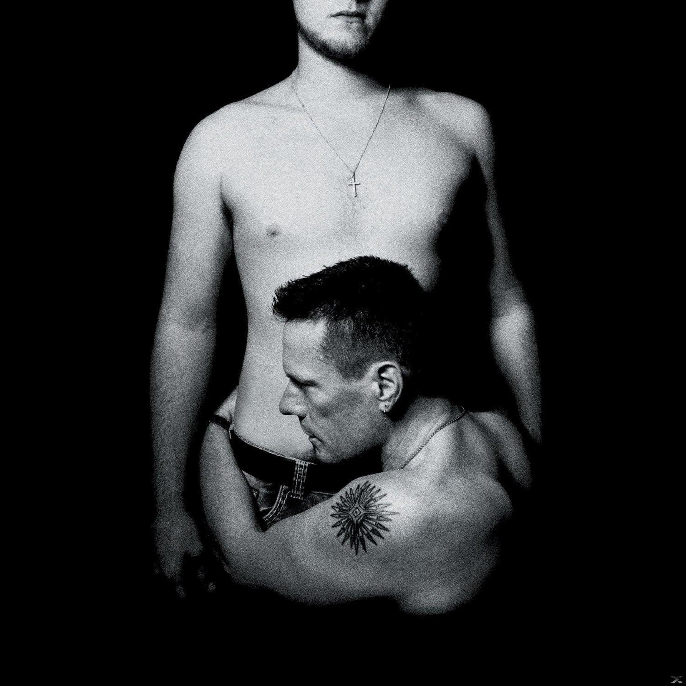 Twee mannen zonder shirt die elkaar omarmen, een met een tatoeage, op een zwarte achtergrond.