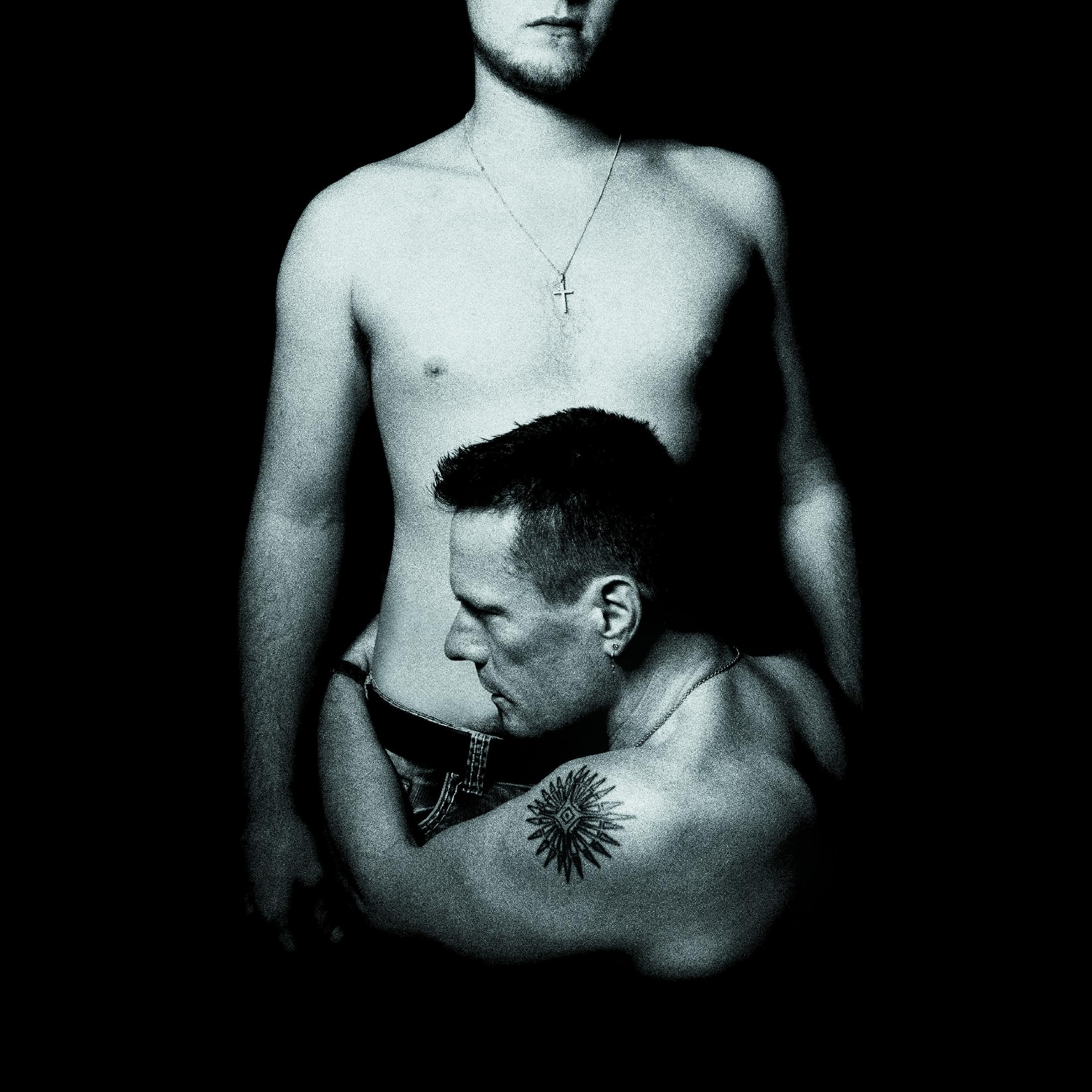 Twee mannen zonder shirt die elkaar omhelzen, één met een tatoeage. Zwarte achtergrond.
