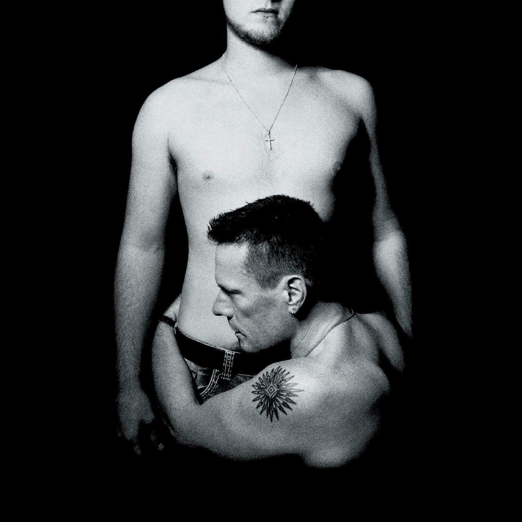 Twee mannen zonder shirt die elkaar omarmen, een met een tatoeage. Zwart-wit afbeelding.