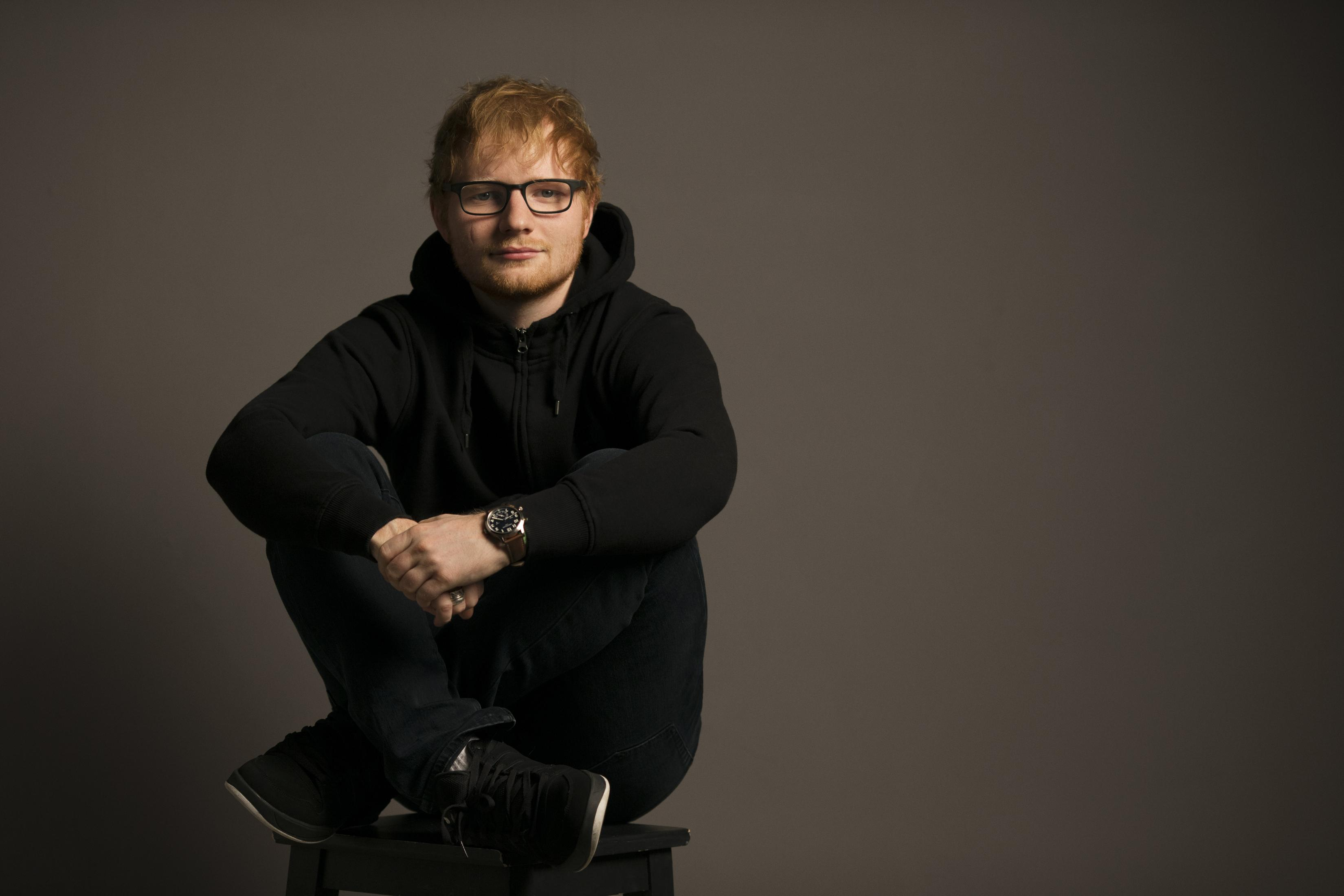 Ed Sheeran zit op een kruk, draagt een bril en een hoodie. Grijze achtergrond.