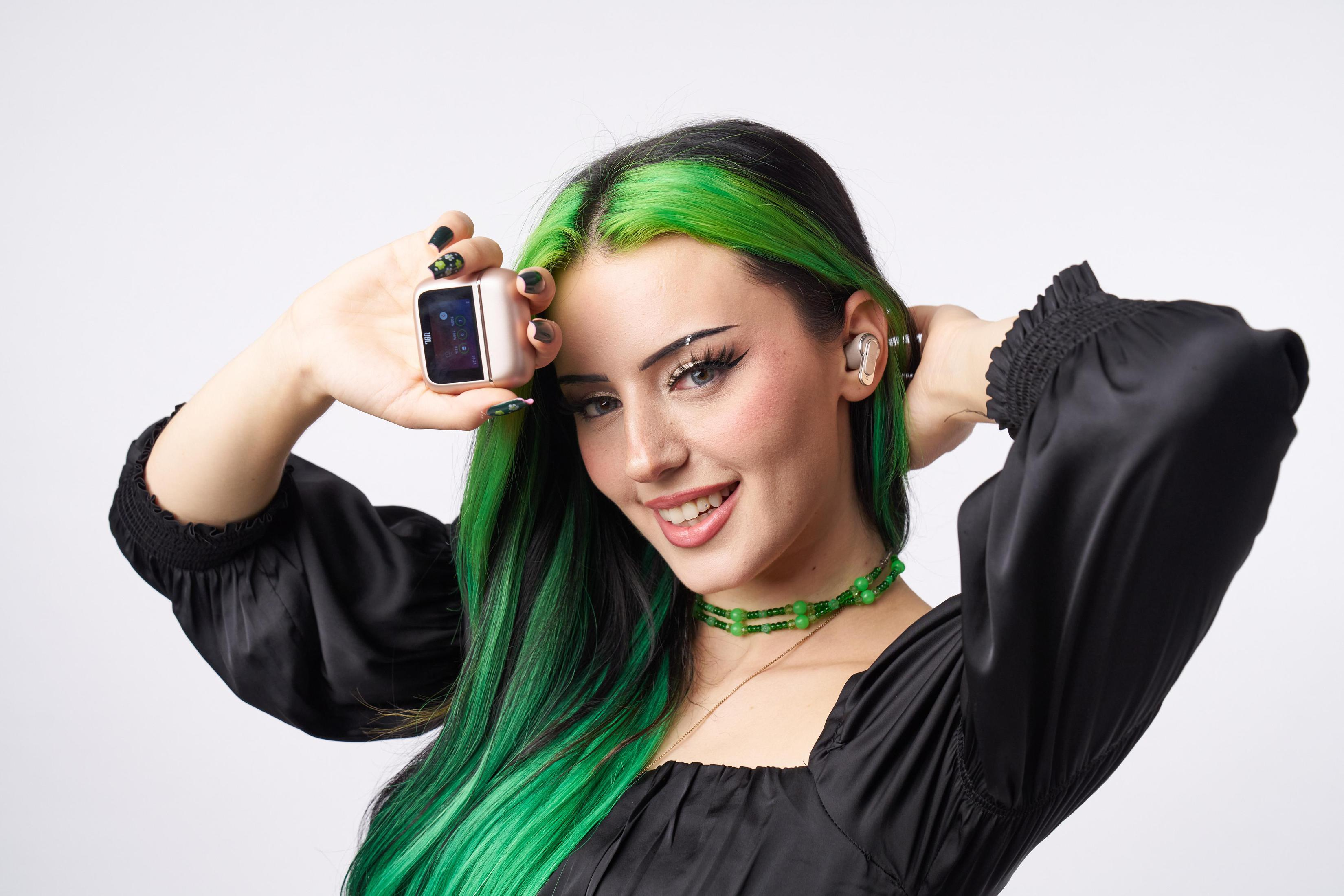 Uśmiechnięta kobieta z zielonymi włosami trzyma smartwatch i słuchawki.