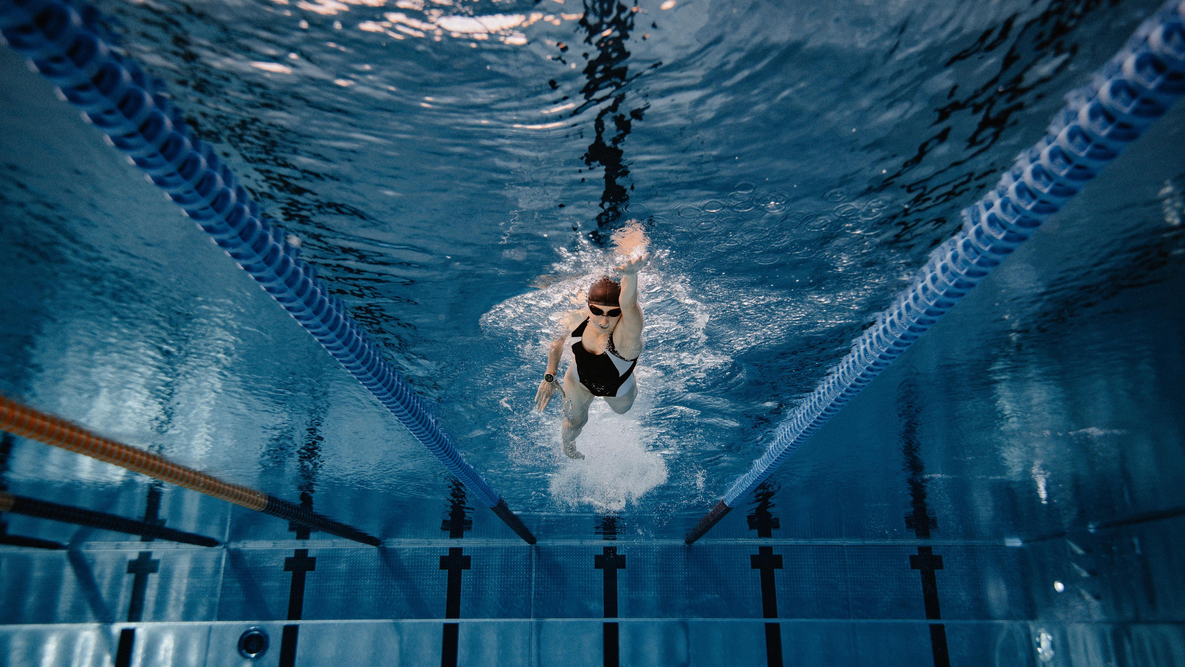 Un nuotatore in piscina visto dal basso. Acqua blu e corsie. Il nuotatore è al centro.
