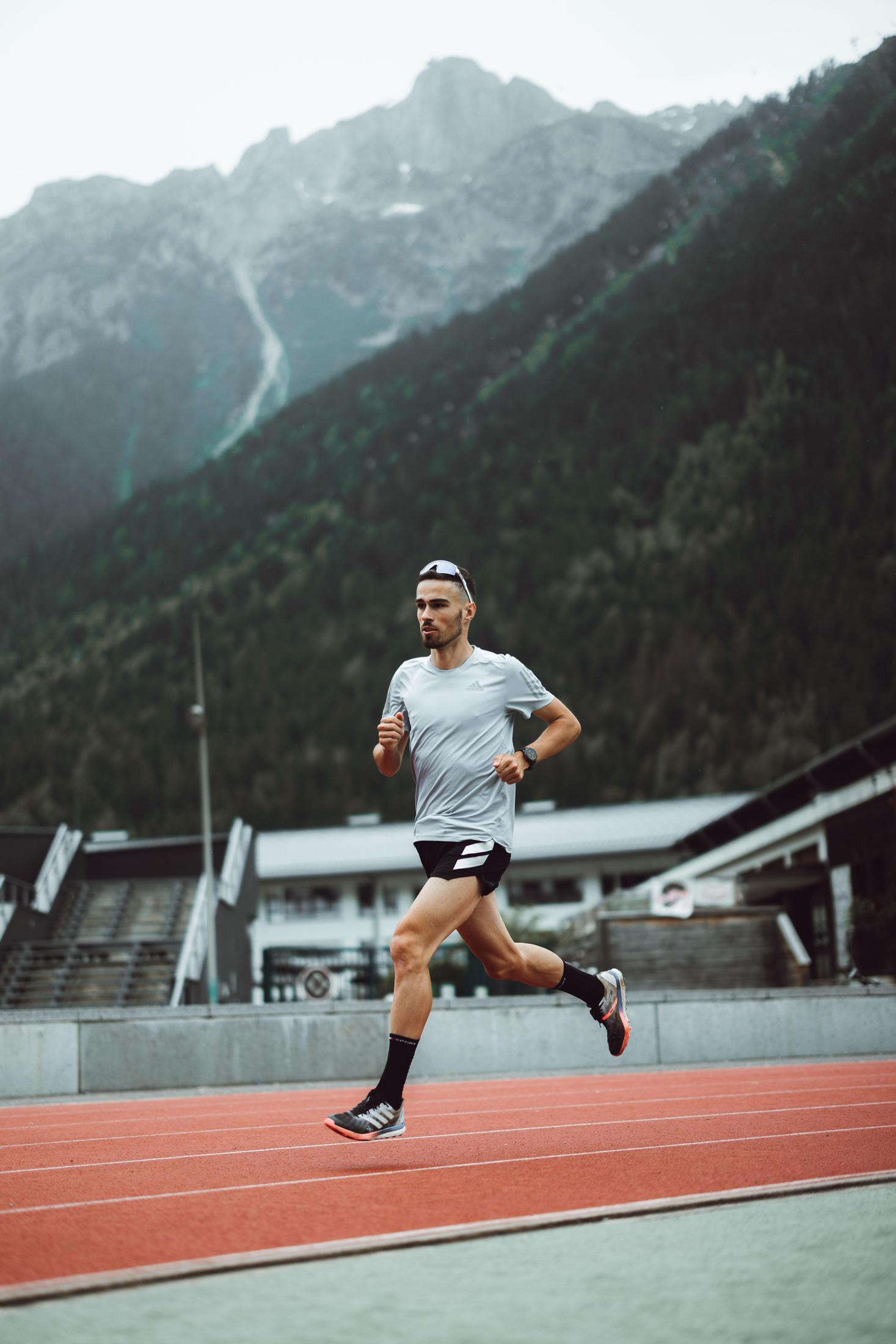 Uomo che corre su pista rossa, montagne sullo sfondo, nuvoloso.