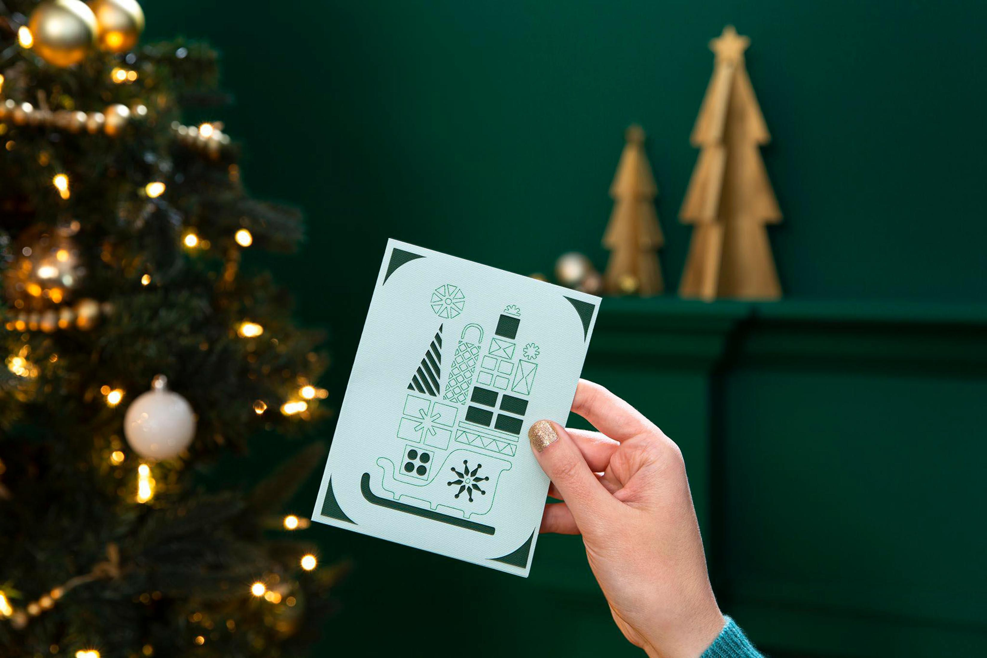 Hand hält Weihnachtskarte mit Geschenken und Schlitten-Design vor Weihnachtsbaum und grüner Wand.