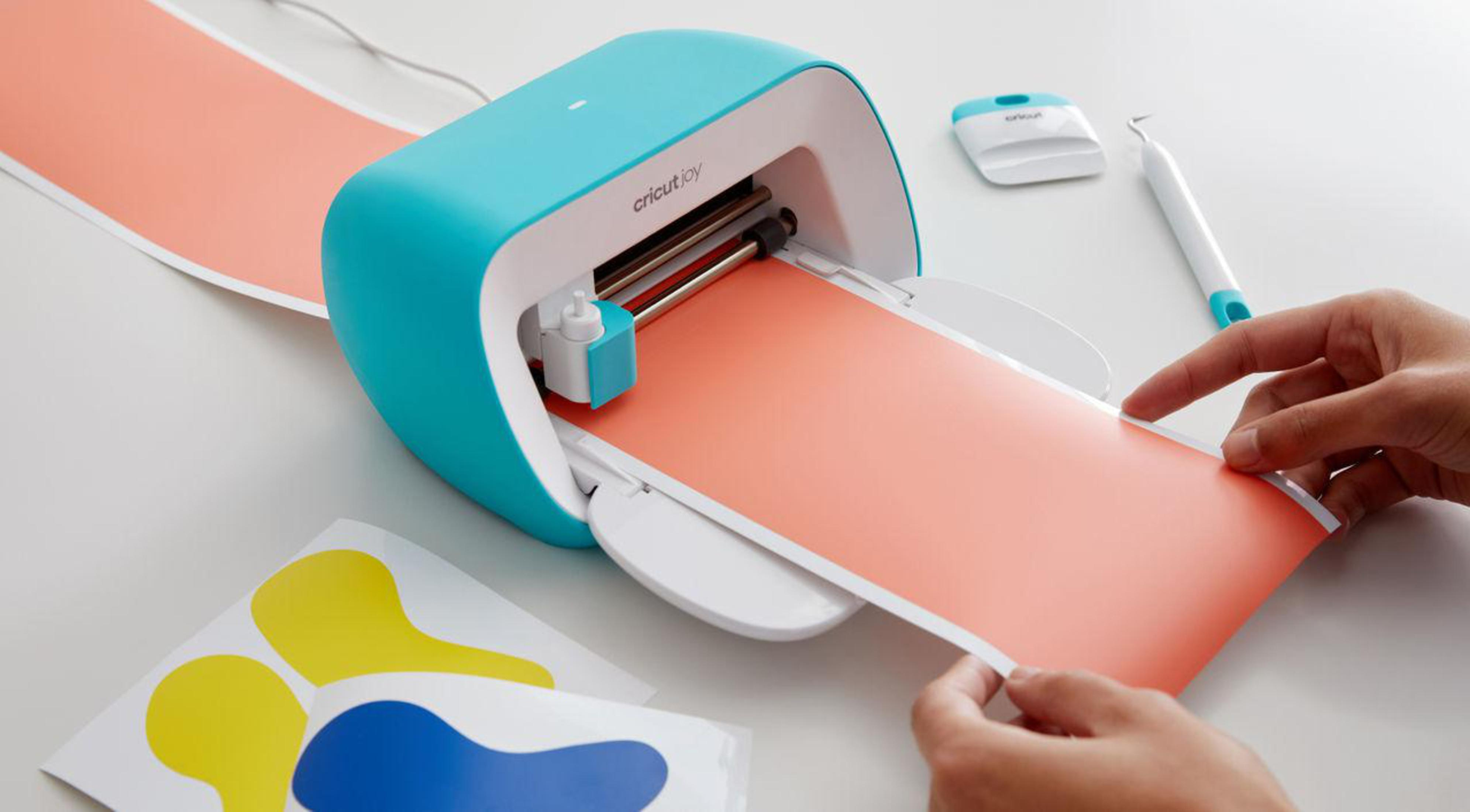 Eine Cricut Joy Maschine, durch die orangefarbenes Material geführt wird. Daneben ein kleiner Cutter und Stift.