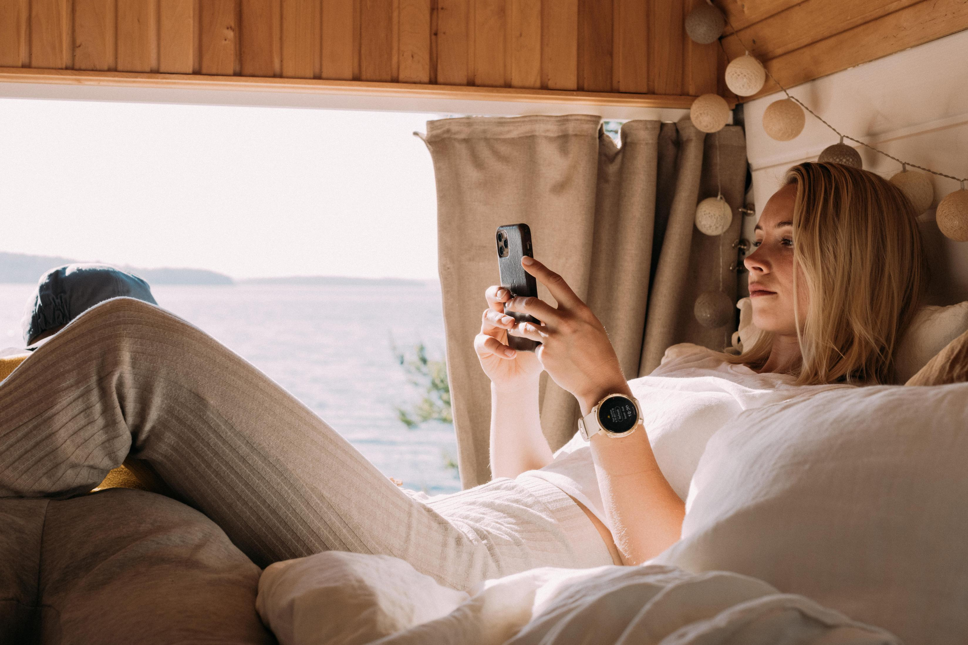 Kobieta w łóżku korzystająca ze smartfona. Zegarek na nadgarstku. Tłem jest okno.