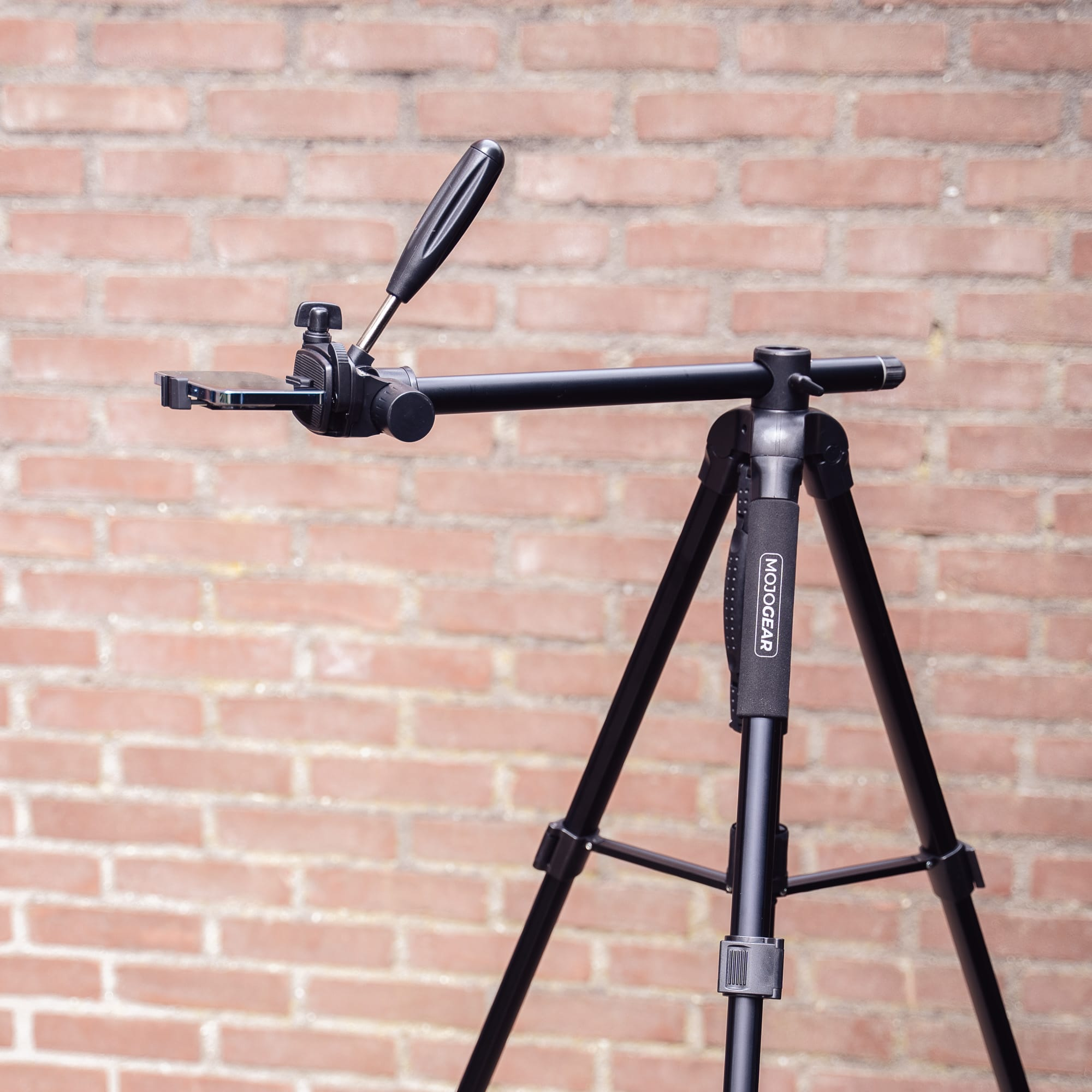 A black tripod with a phone holder attached. The holder is holding a phone. A brick wall is in the background.
