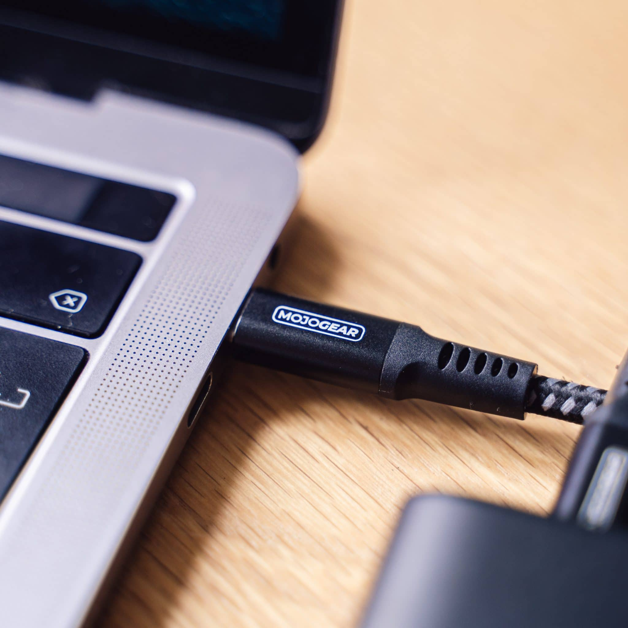 Een zwarte MOJOGEAR USB-C-kabel aangesloten op een laptop. Kabel is zwart met grijze accenten. Houten tafel.