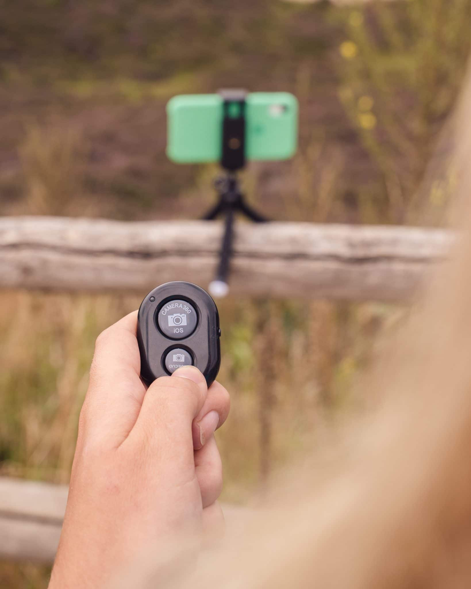 A hand holds a remote for a phone on a tripod. A green phone is mounted on the tripod, in a field.