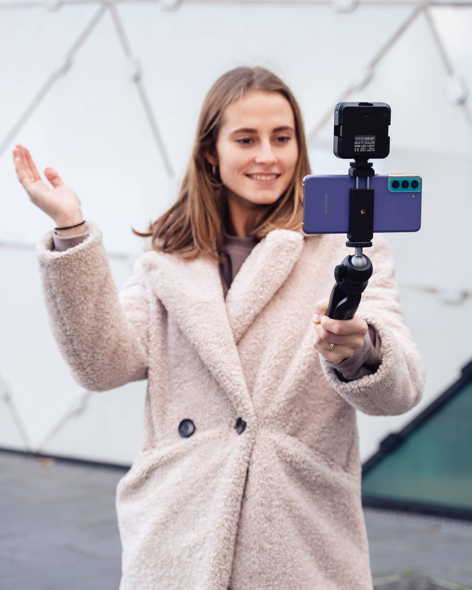 Woman vlogging with a phone on a tripod, wearing coat. Background is a white building.