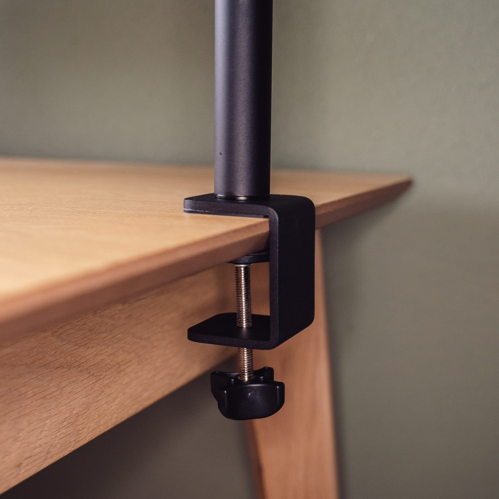 Zwarte monitorstandaardklem, die een zwarte paal vasthoudt. Het is bevestigd aan een houten tafel. De achtergrond is groen.