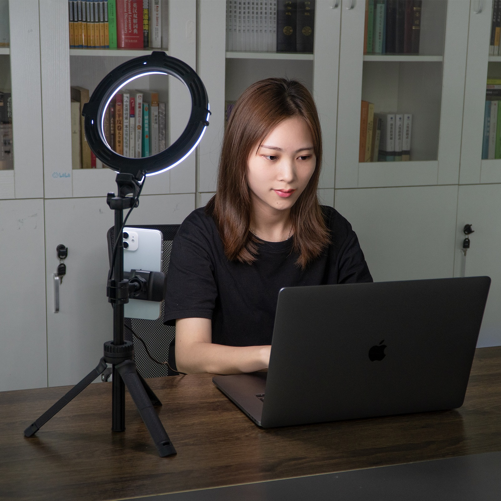 Woman using a laptop with ring light and phone holder on desk. Bookshelf in the background.