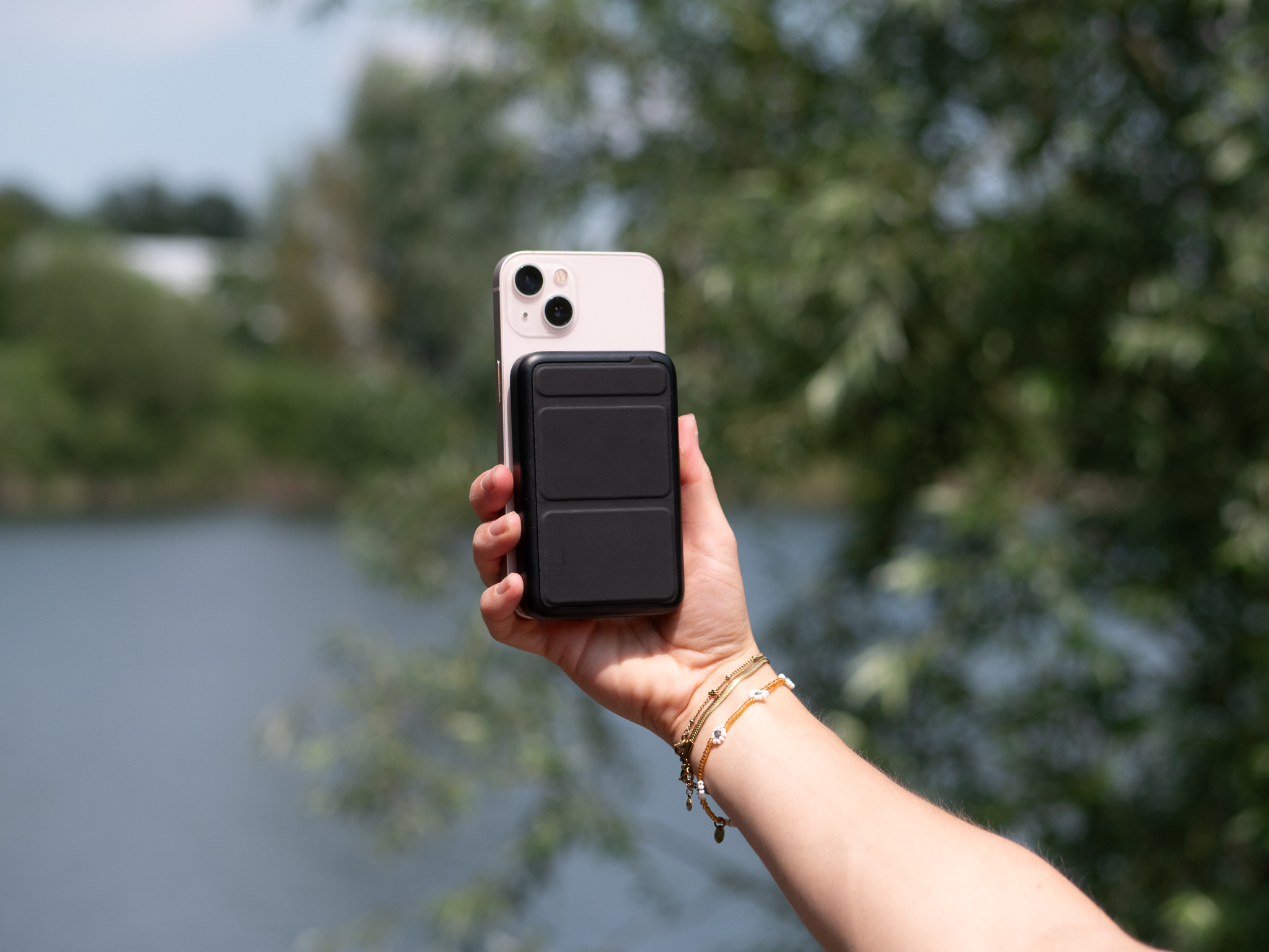 Een hand die een telefoon vasthoudt met een zwarte oplader eraan. De achtergrond is een groen veld en water.