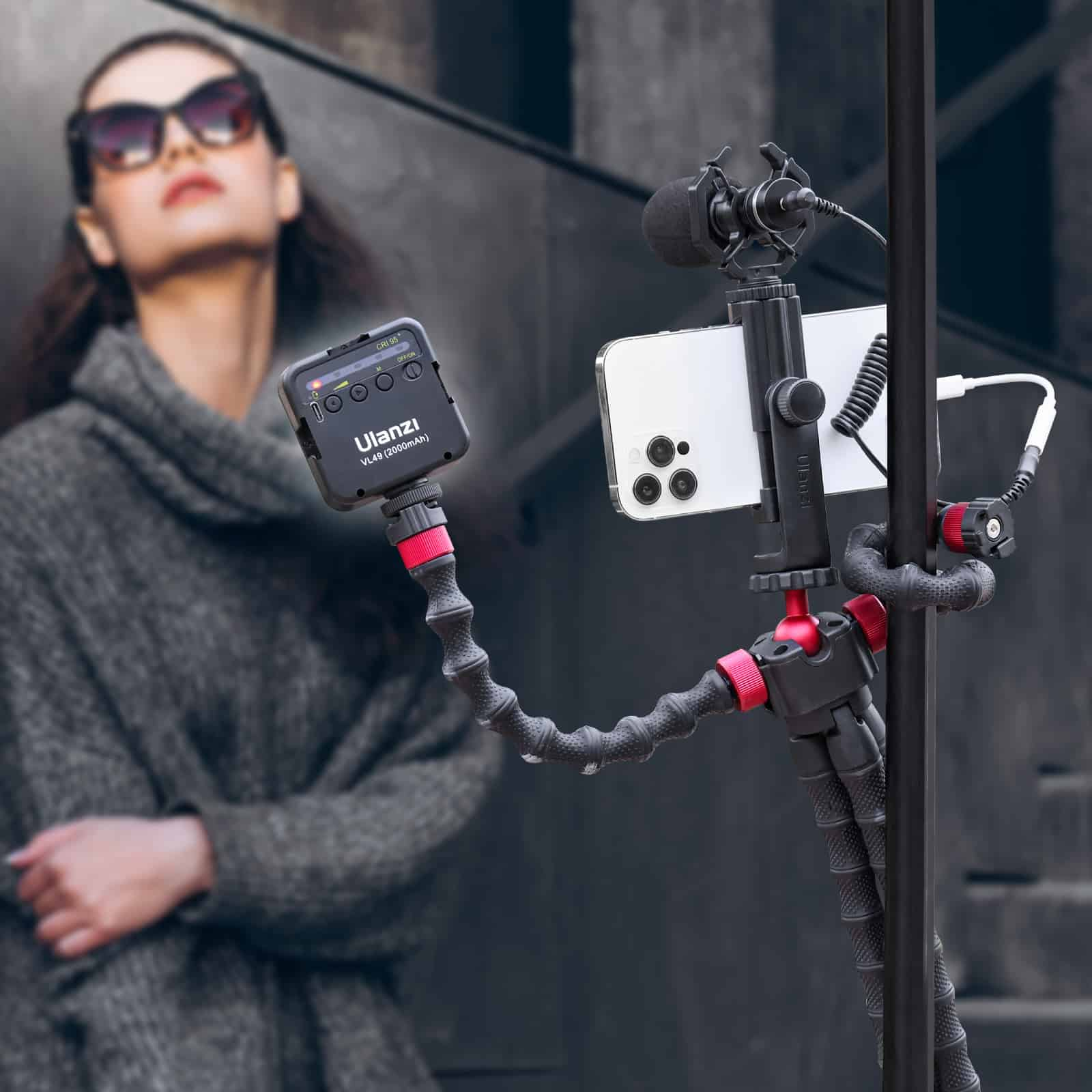 A woman in sunglasses holds a phone with a microphone and light attached to a flexible tripod.