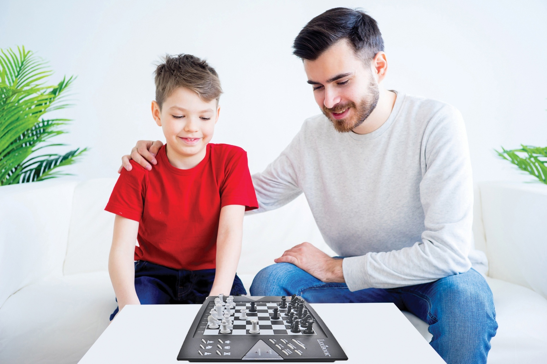Padre e hijo jugando al ajedrez. Padre a la derecha, hijo a la izquierda, y el tablero de ajedrez al frente. Sofá blanco.