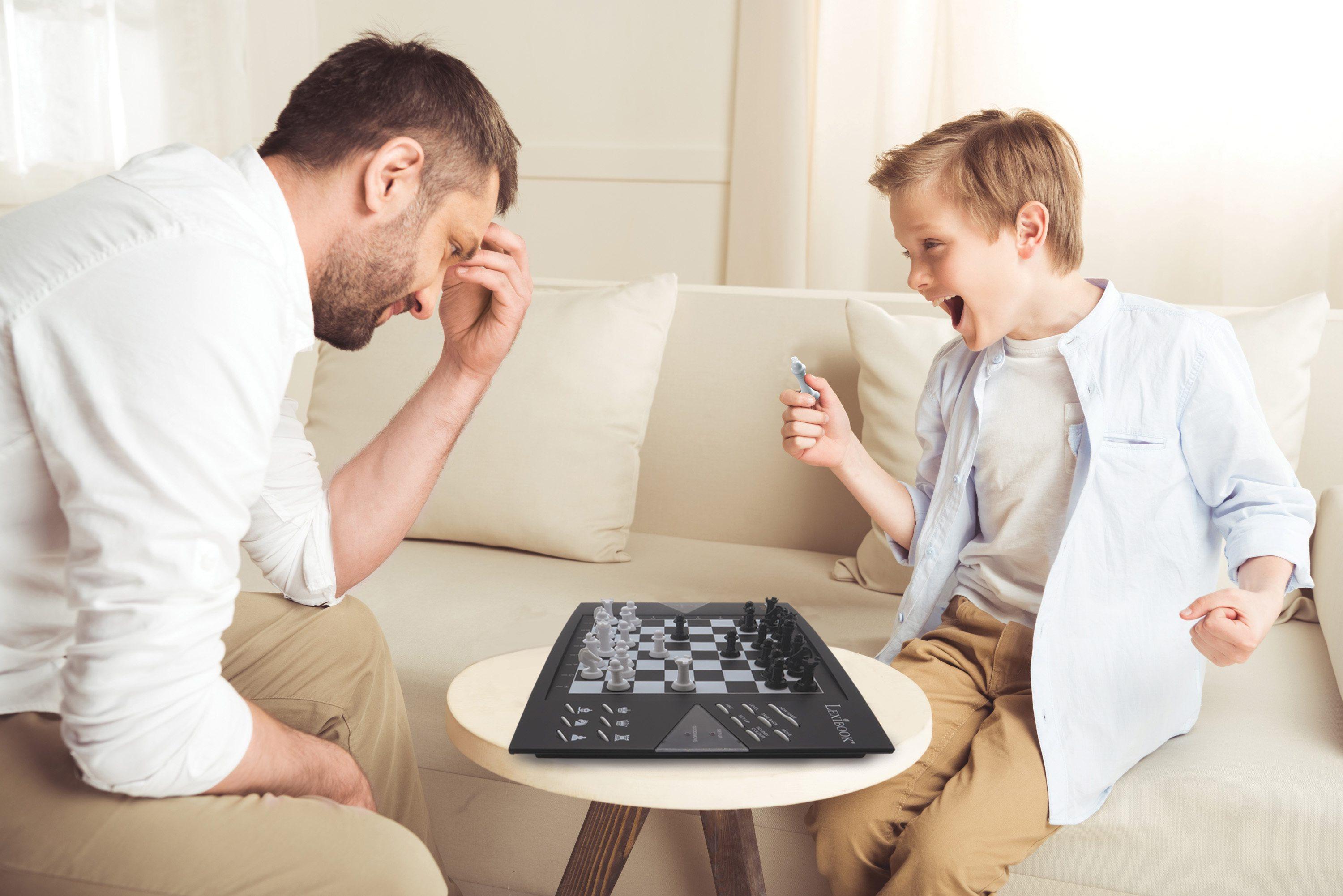 Padre e hijo jugando ajedrez. El padre está en shock, el hijo emocionado. Tablero de ajedrez al frente, sofá blanco al fondo.