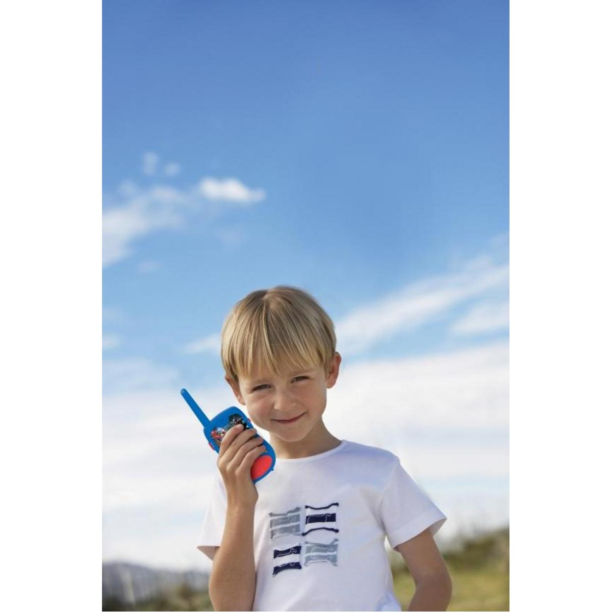 Un niño sosteniendo un walkie-talkie azul, sonriendo frente a un cielo azul y nubes.