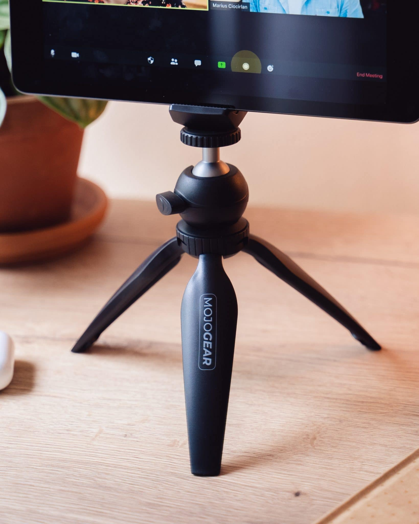 Een klein zwart statief met een tablet waarop een videogesprek wordt getoond. Het statief staat op een houten bureau.