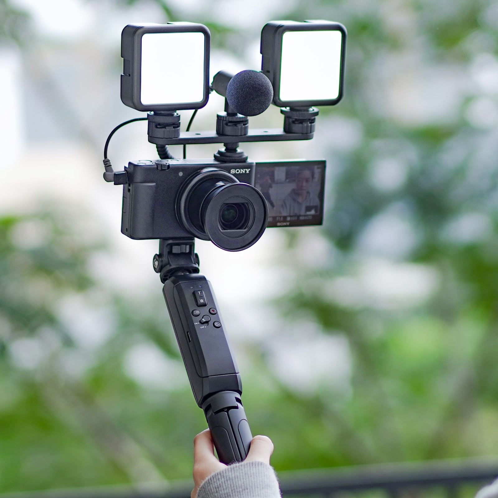 A camera setup with lights and a microphone, held by a hand, against a blurred background.