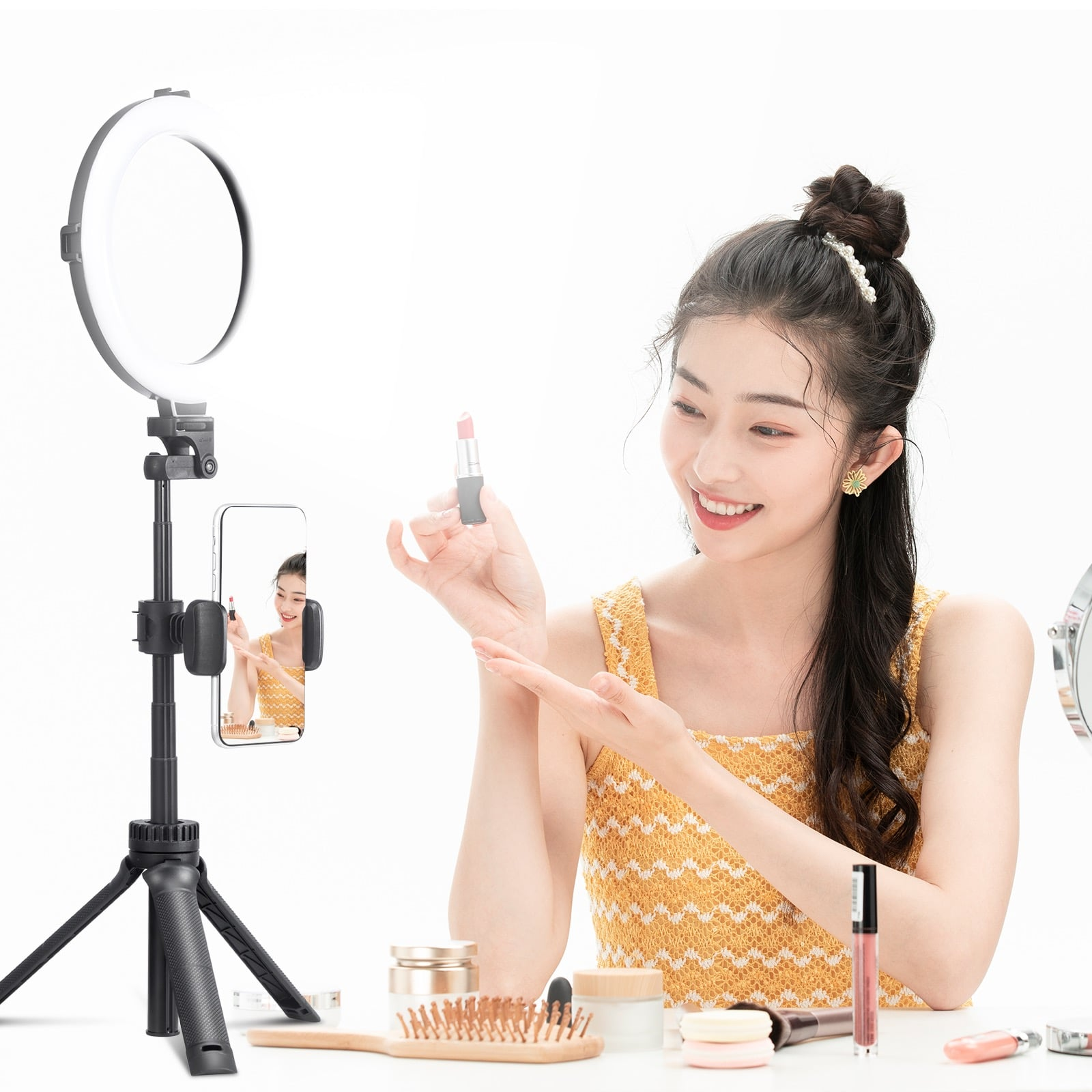 Woman holding lipstick, filming with phone on tripod with ring light. Makeup on the table.