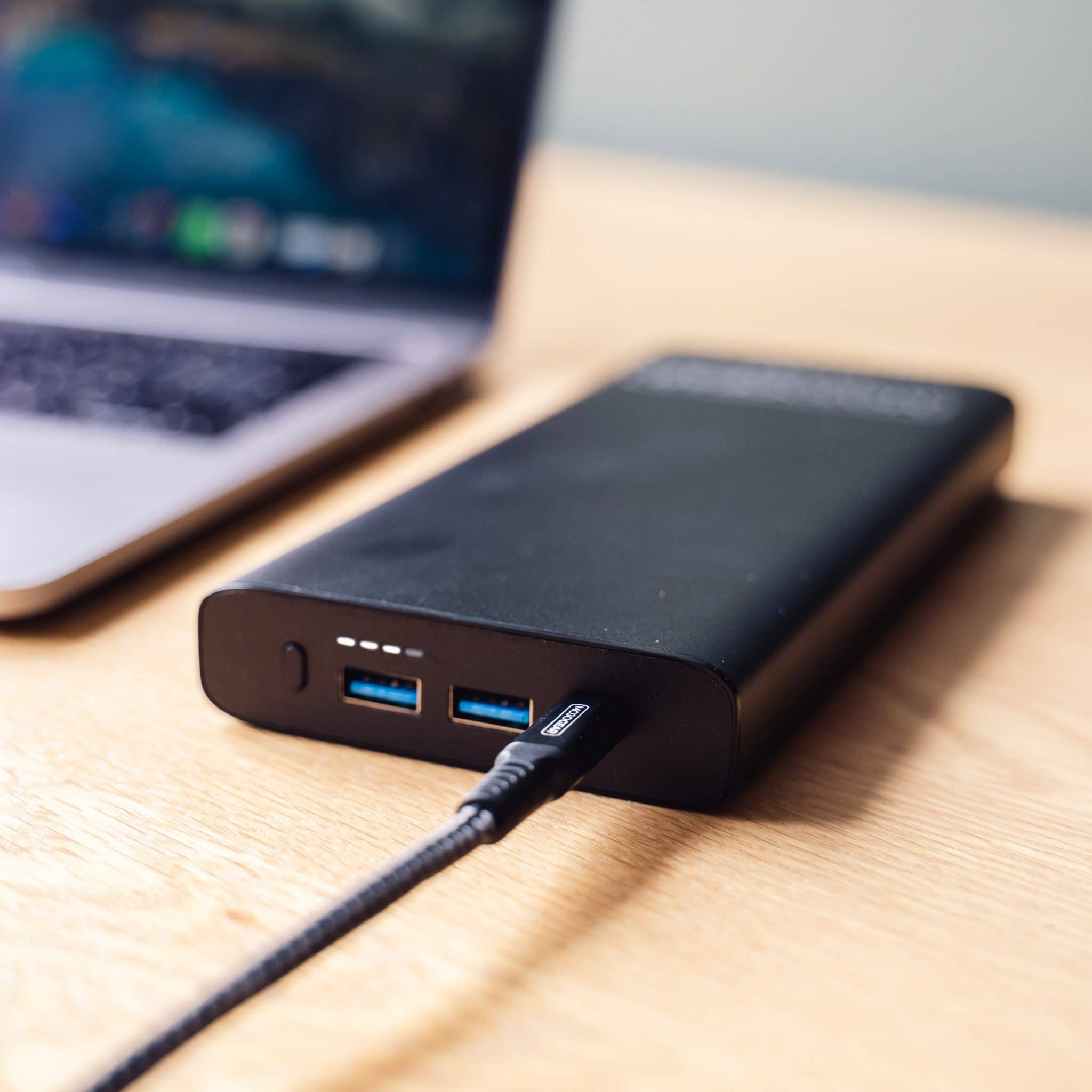 Zwarte powerbank met USB-poorten en een oplaadkabel bij een laptop.