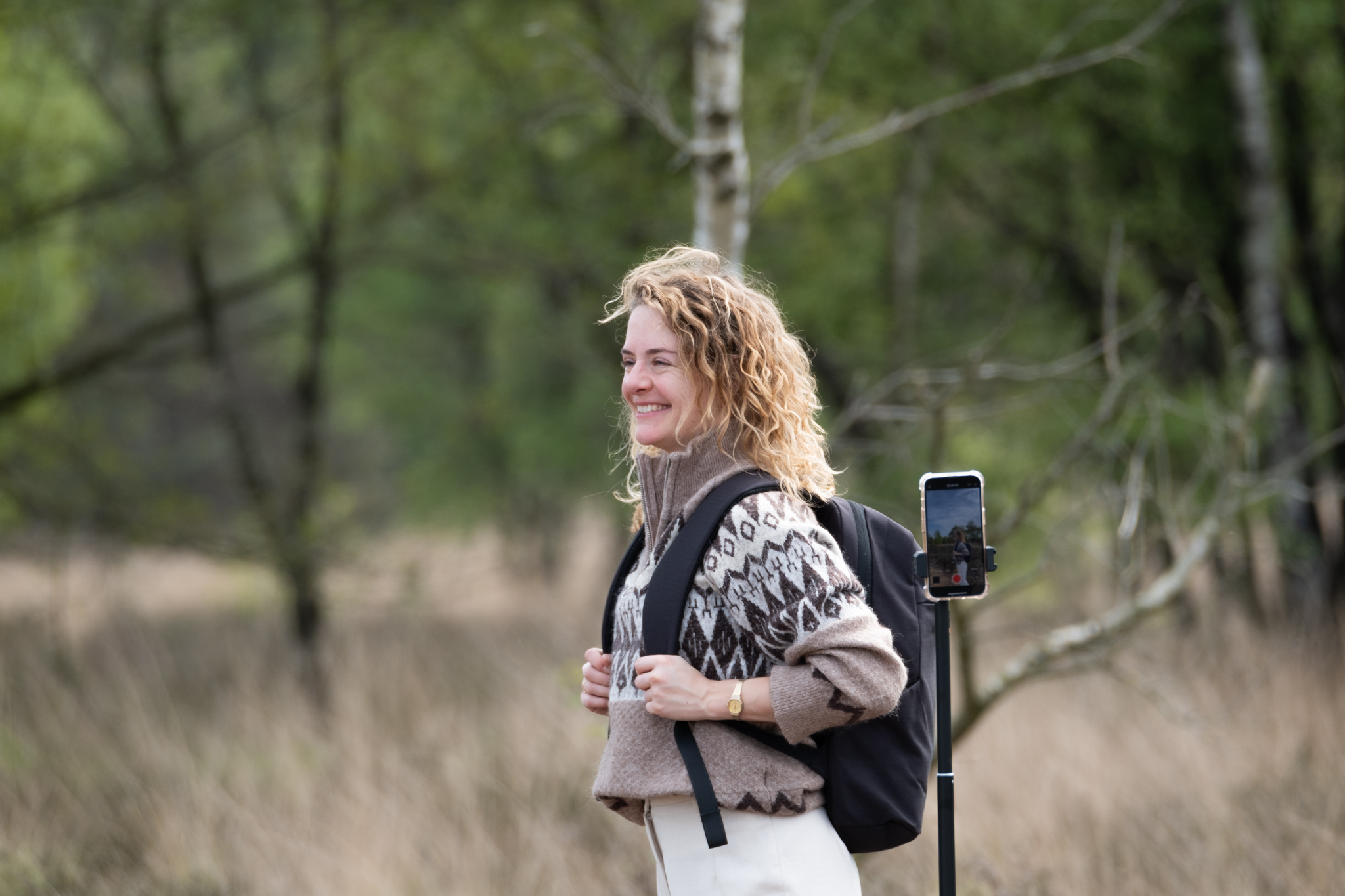 Woman with backpack smiles, holding a phone on a selfie stick, in a green forest with trees.