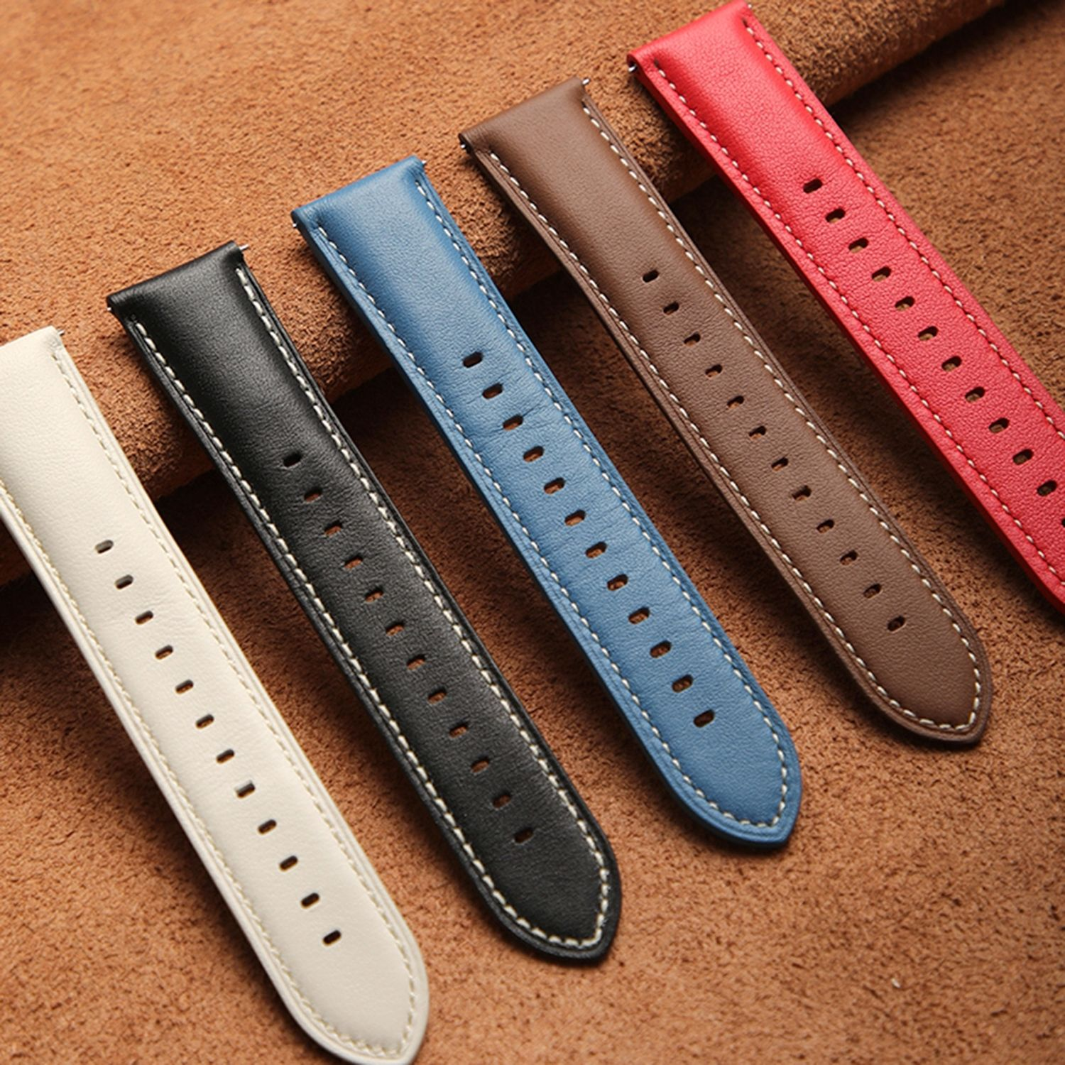Fünf Leder-Uhrenarmbänder in verschiedenen Farben: Weiß, Schwarz, Blau, Braun und Rot, angeordnet auf einer strukturierten Oberfläche.
