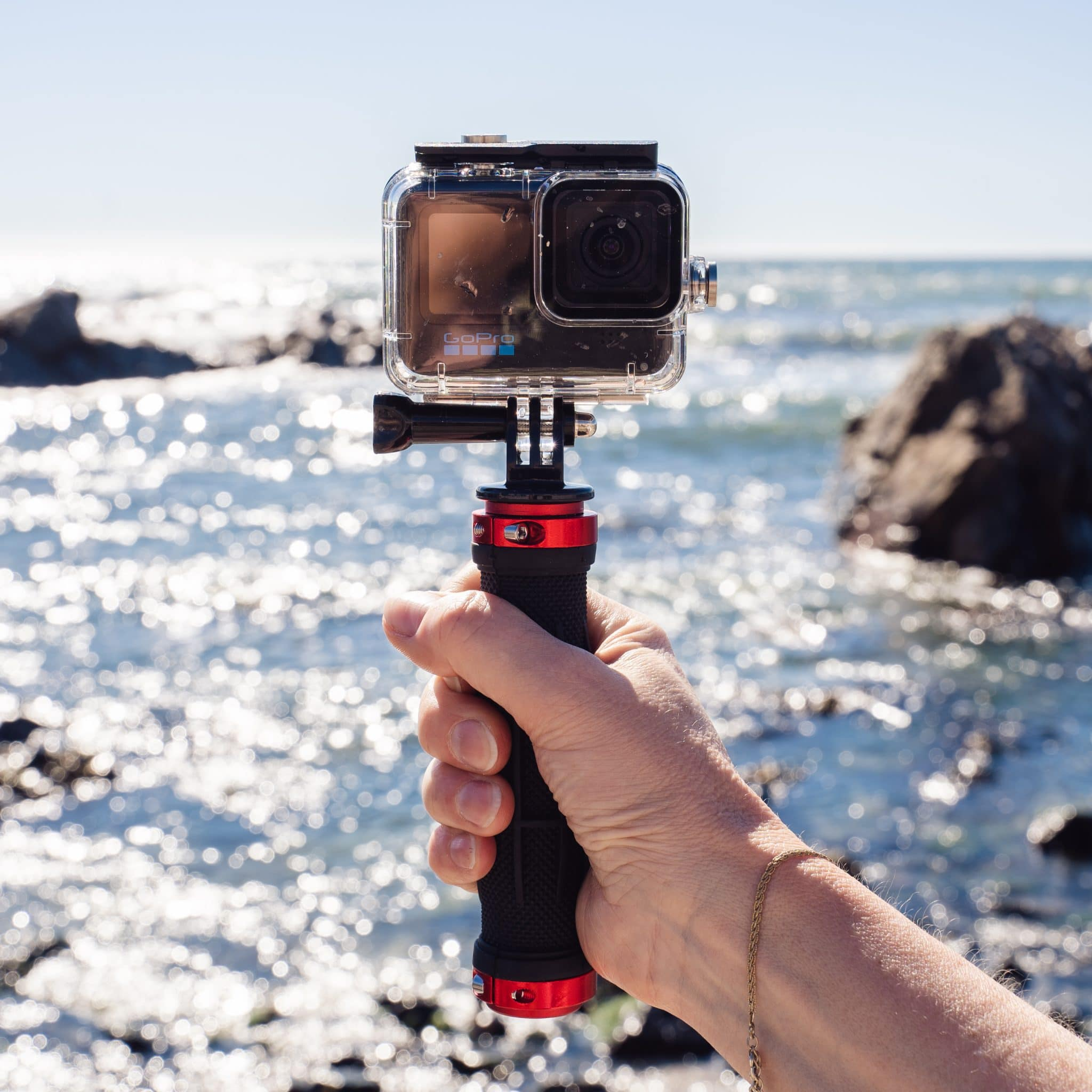 De hand van een persoon houdt een GoPro-camera vast in een waterdichte behuizing. De achtergrond is de zee.