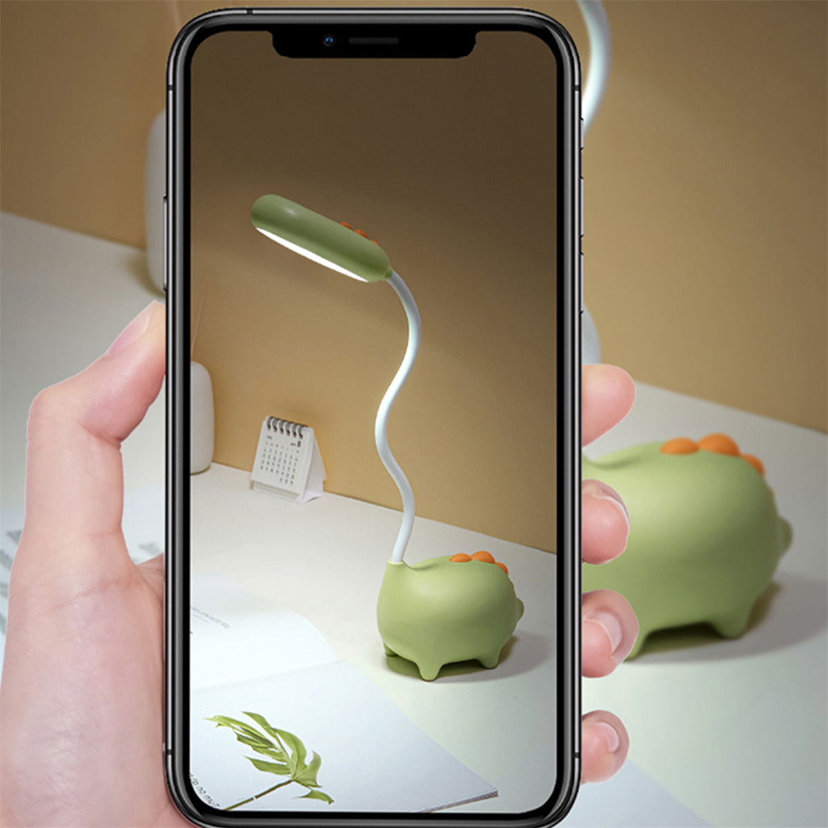 Eine Hand hält ein Telefon, das eine grüne Lampe in Dinosaurierform zeigt, auf einem Schreibtisch.