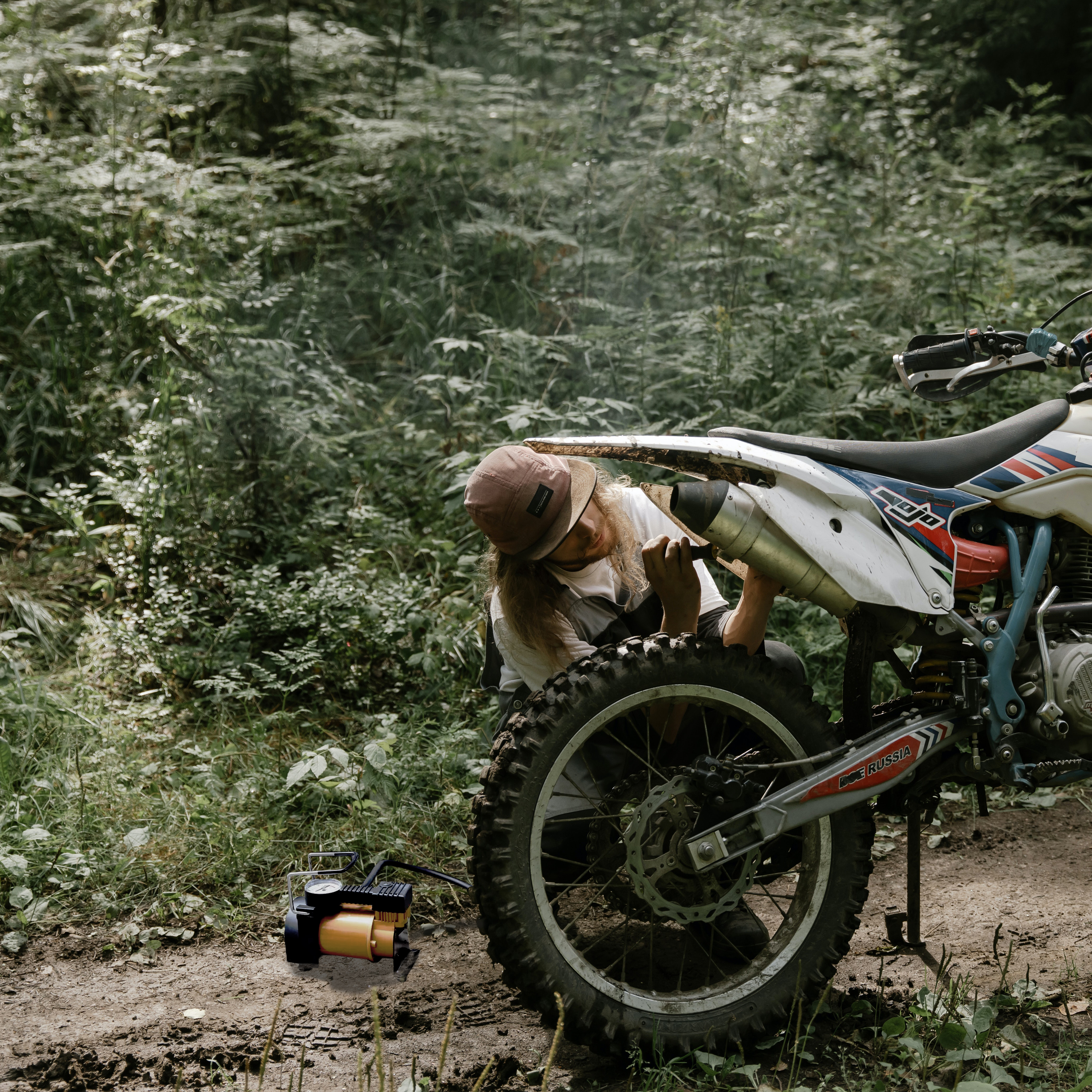 Una persona lavora su uno pneumatico di una moto in un ambiente forestale. Una pompa è vicina. Fogliame verde sullo sfondo.