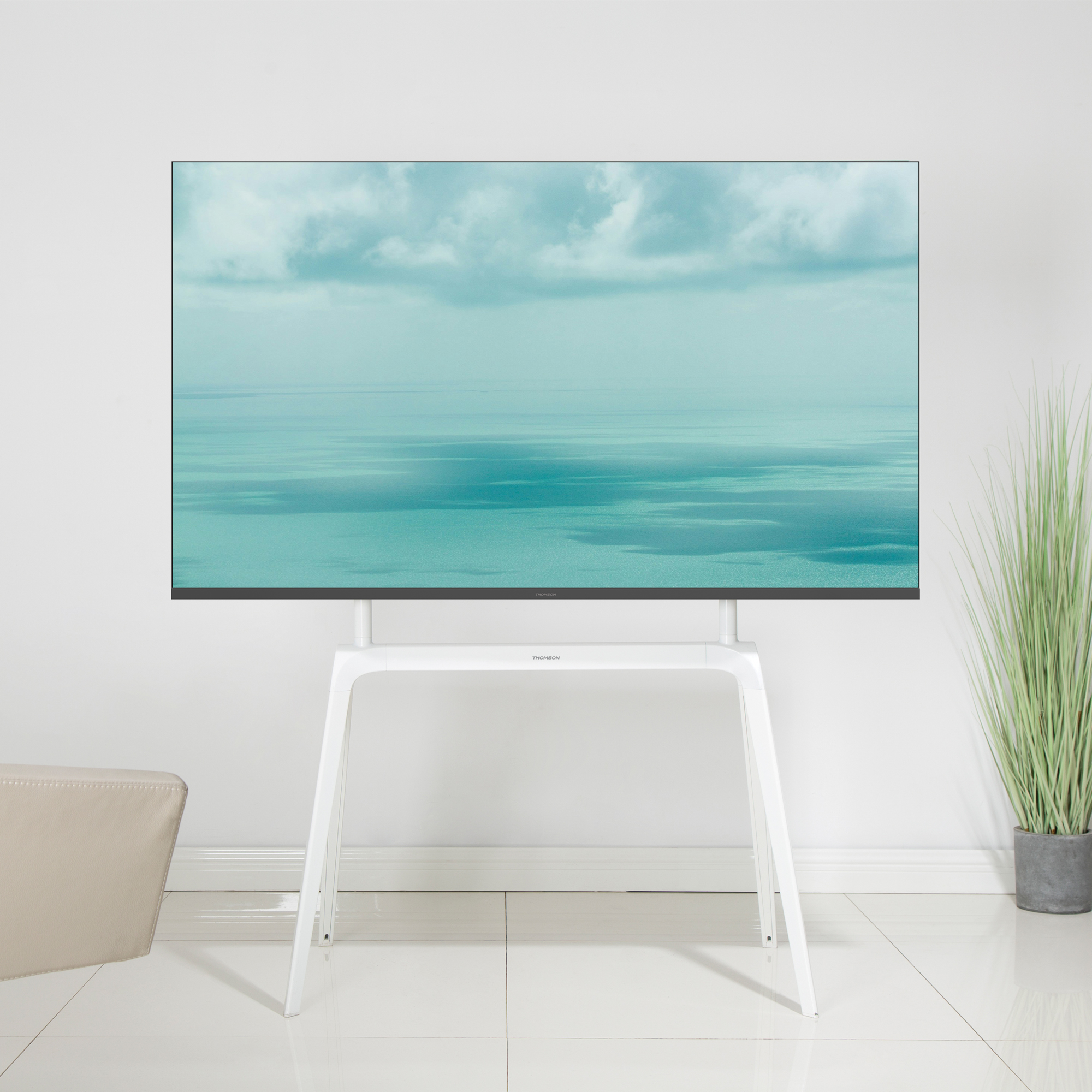 Un grande schermo TV che mostra una scena di mare e cielo turchese, montato su un supporto bianco, in una stanza luminosa.