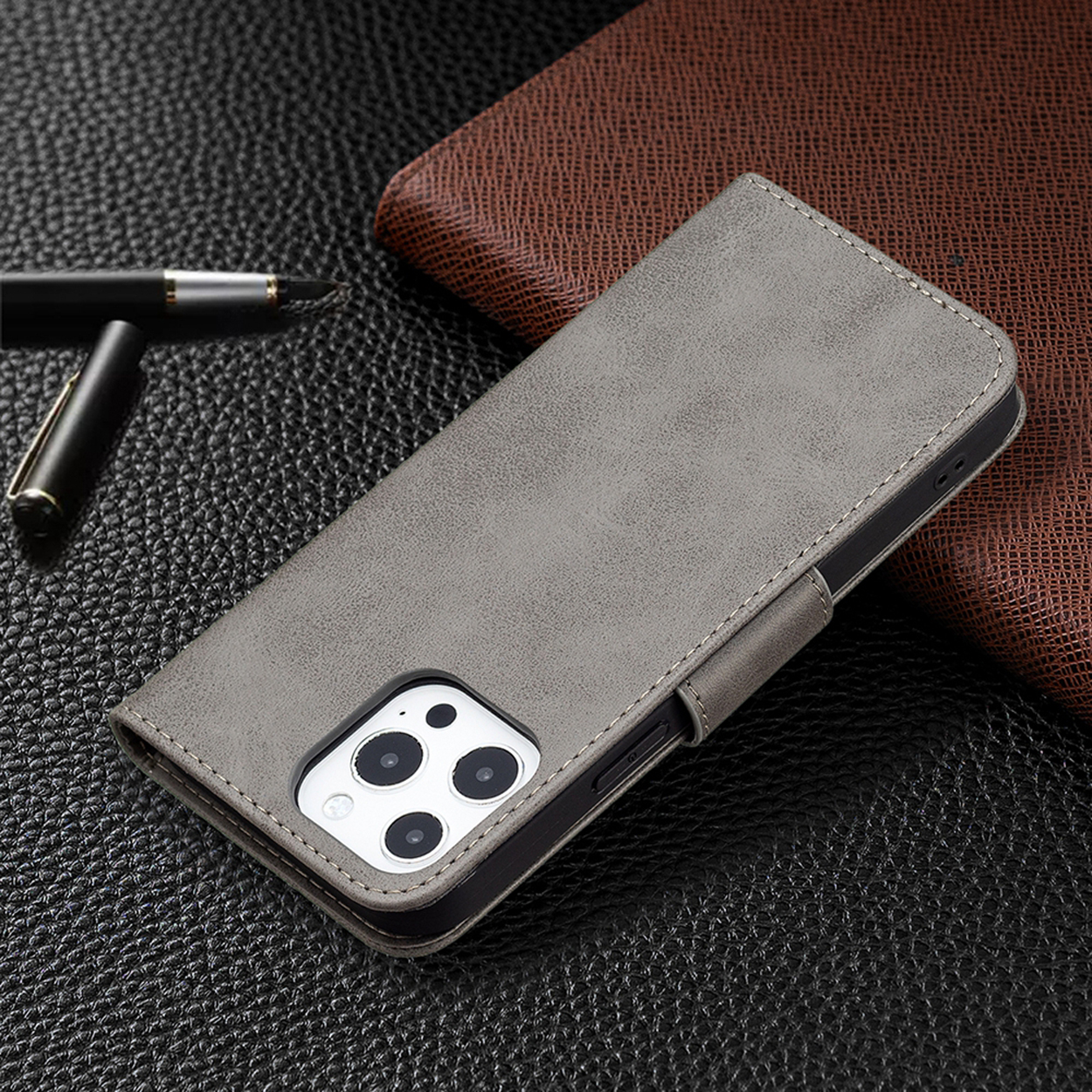 Grijze telefoonhoes met magnetische sluiting, telefoon met camera-uitsparingen, op een getextureerd oppervlak, pen en portemonnee zichtbaar.