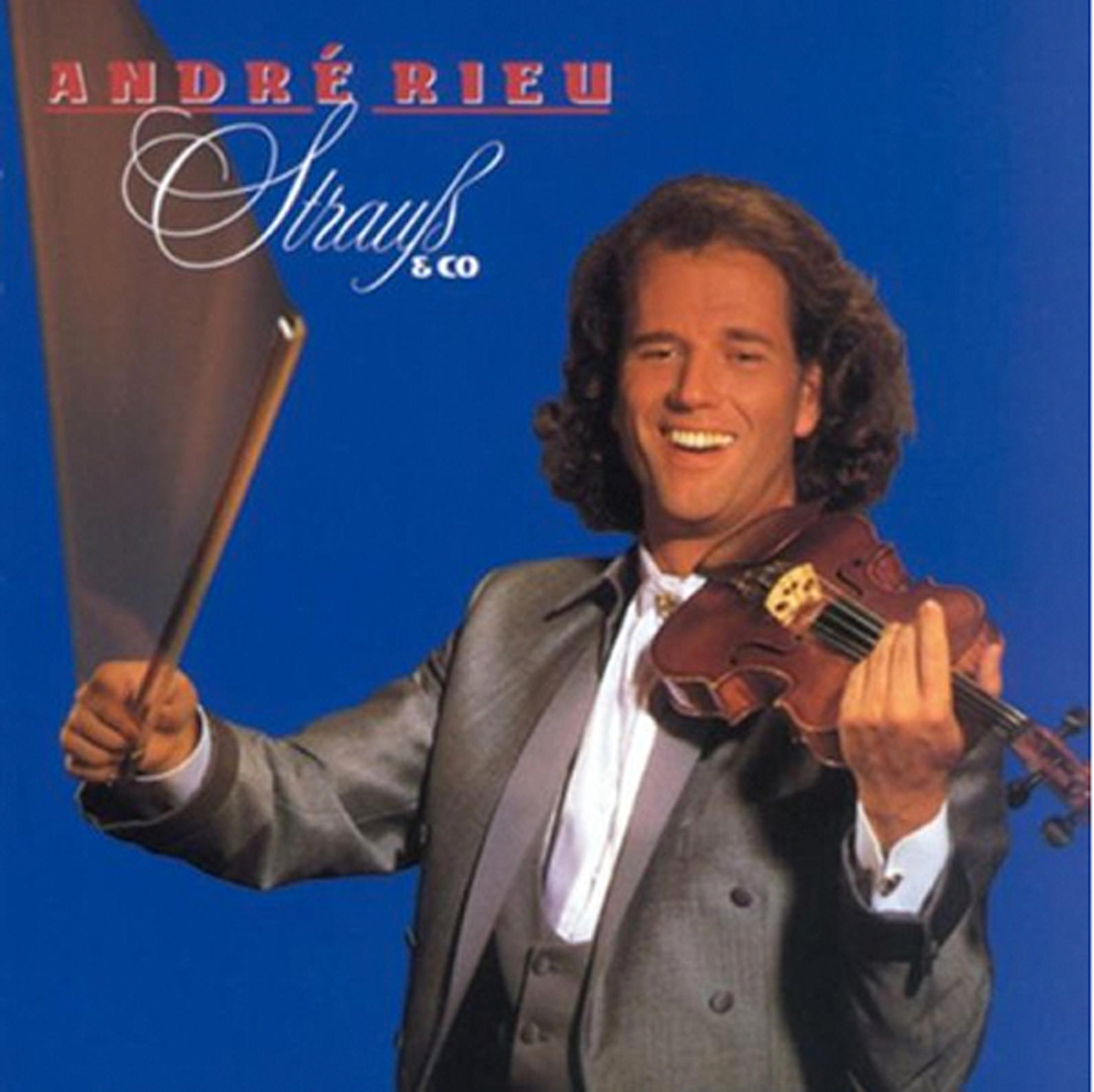 André Rieu souriant, tenant un violon et un archet, en costume gris, sur fond bleu.