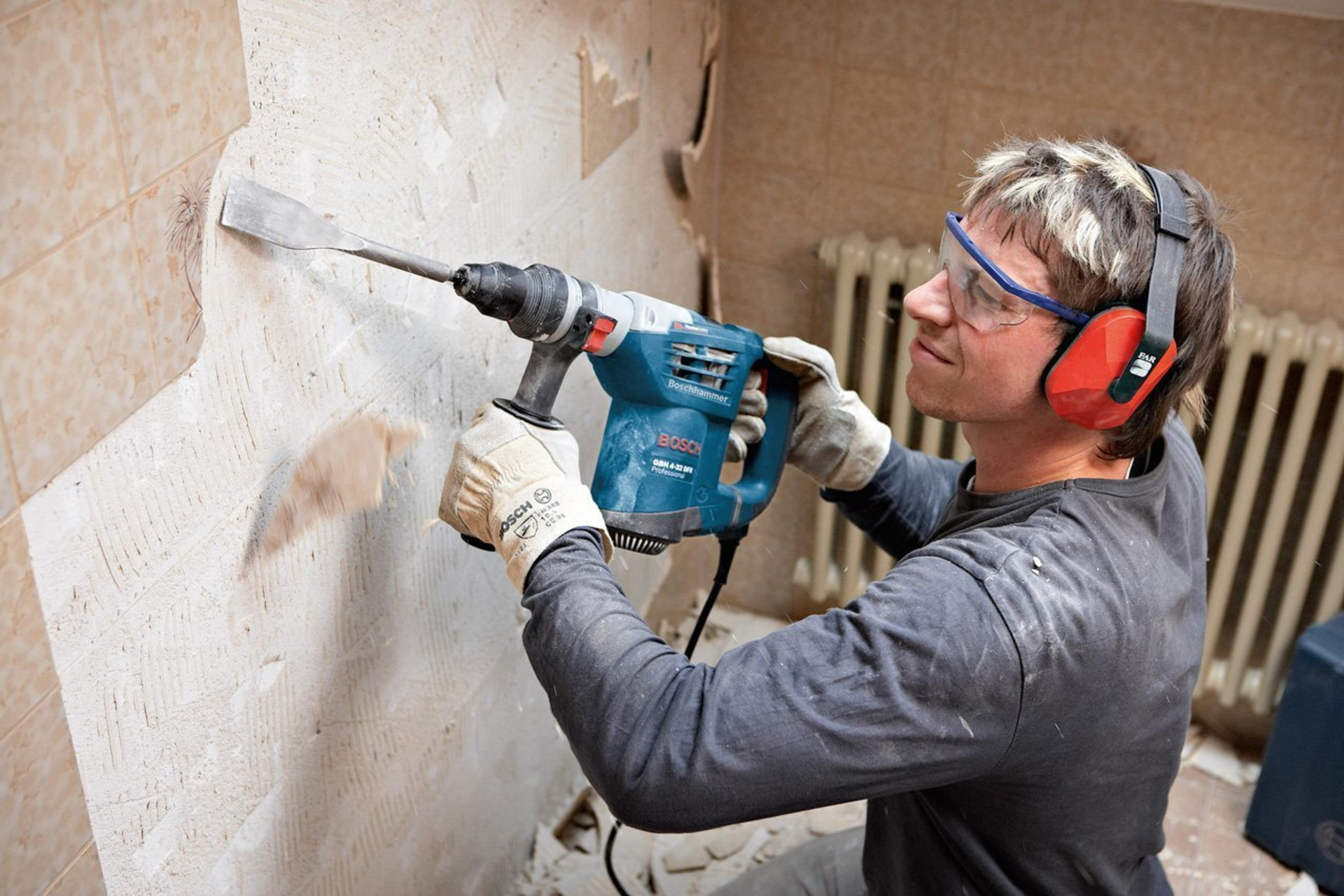 Homme utilisant une perceuse pour enlever des carreaux d'un mur. Il porte des lunettes et une protection auditive.