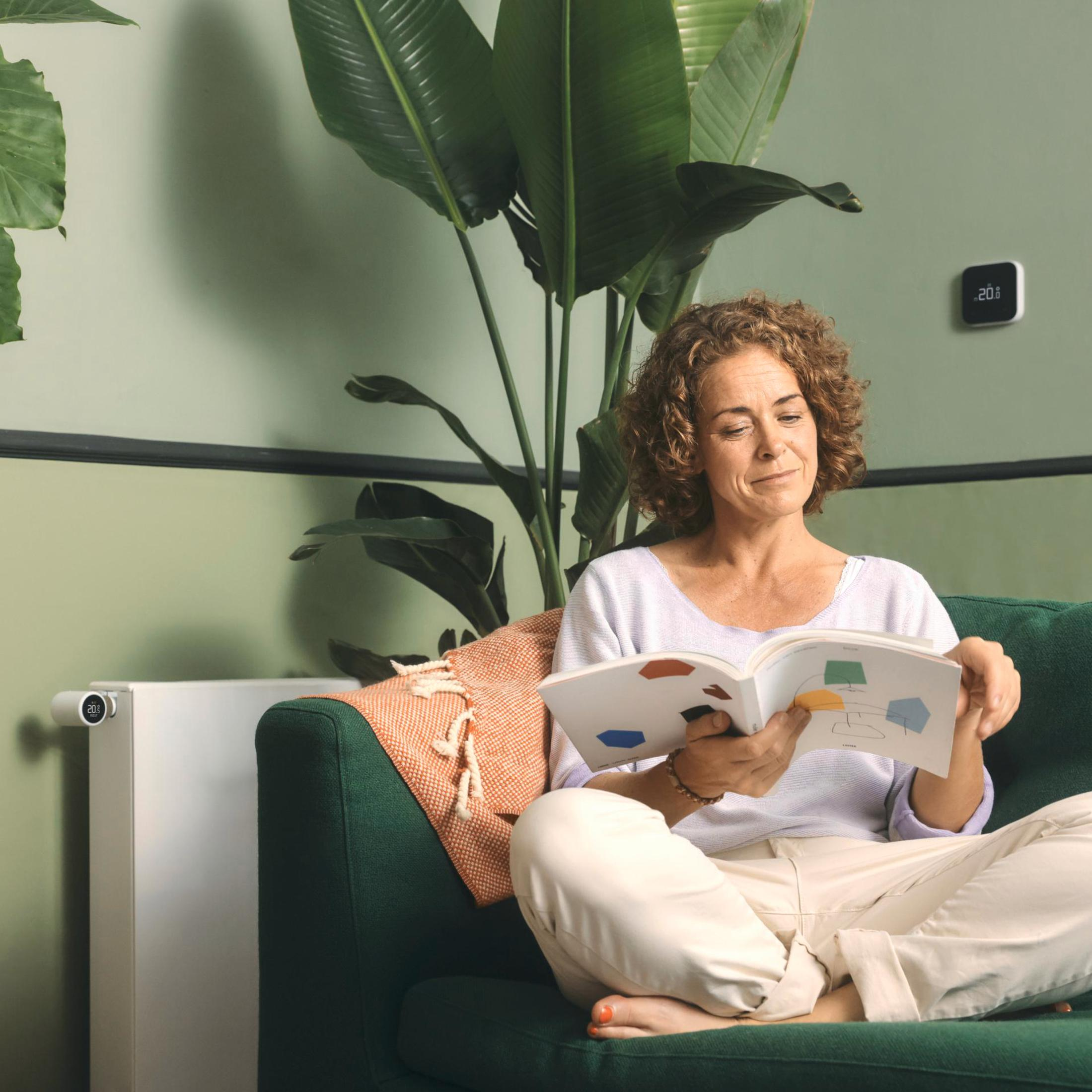 A woman sits on a green couch indoors, reading a book. A plant and a wall-mounted thermostat are visible.
