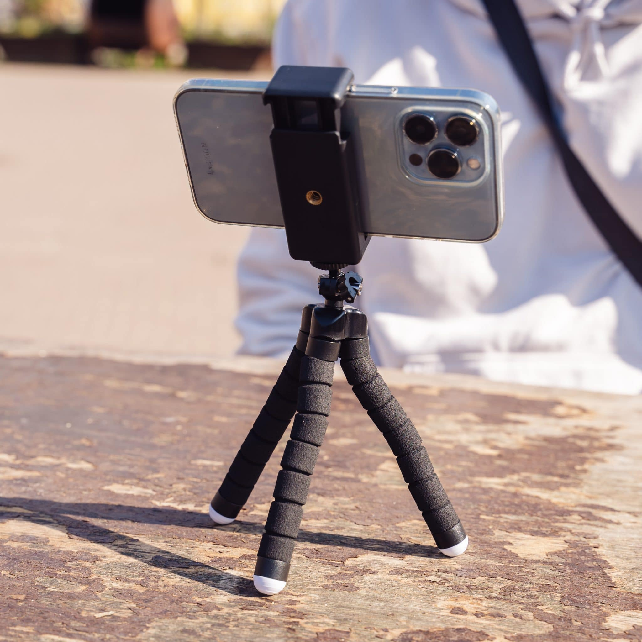 A phone is mounted on a mini tripod. The tripod is on a wooden surface. A person is in the background.