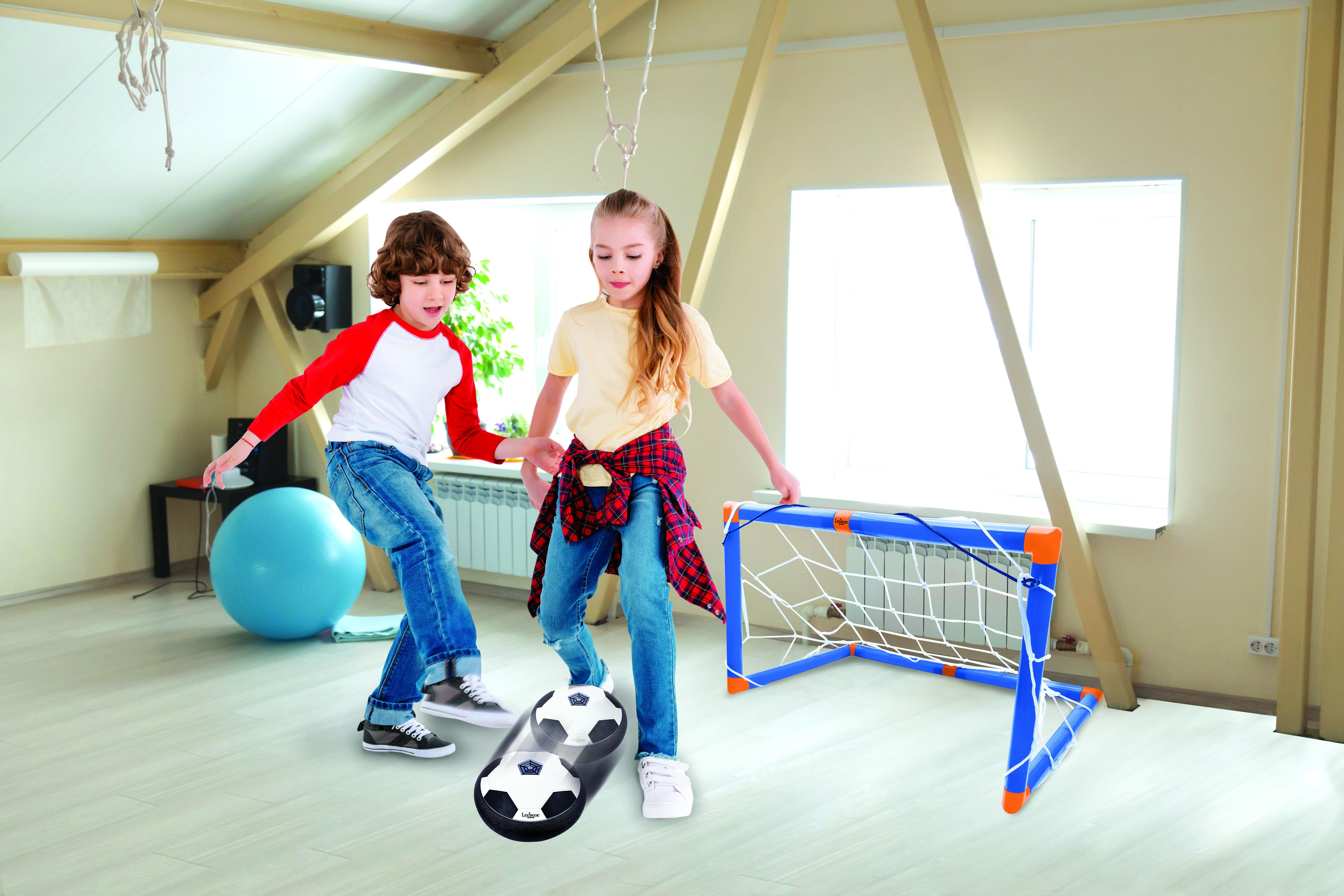 Dos niños jugando al fútbol en una habitación con una portería. Uno patea la pelota. El suelo es de madera clara.