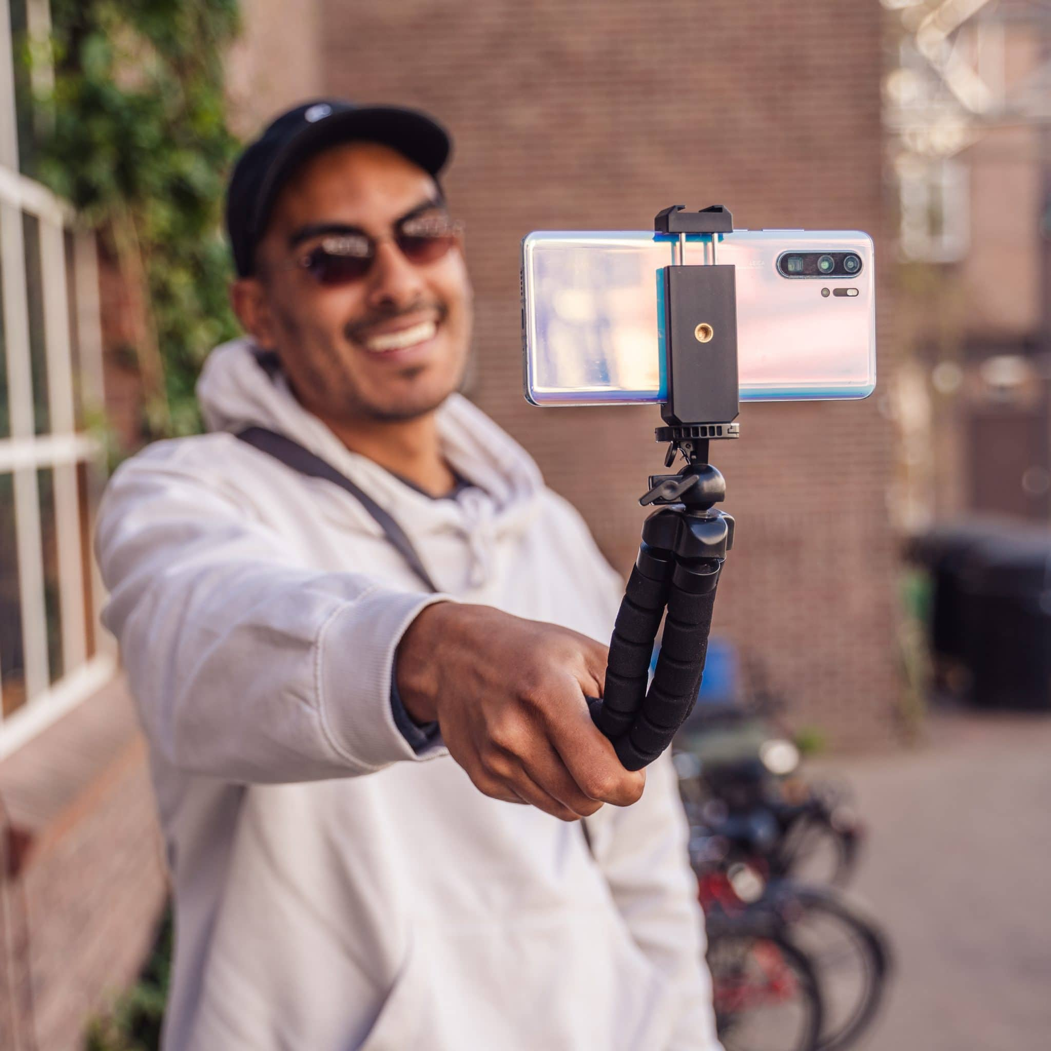 Een lachende man houdt een telefoon op een statief en maakt een selfie. De achtergrond is een straat.