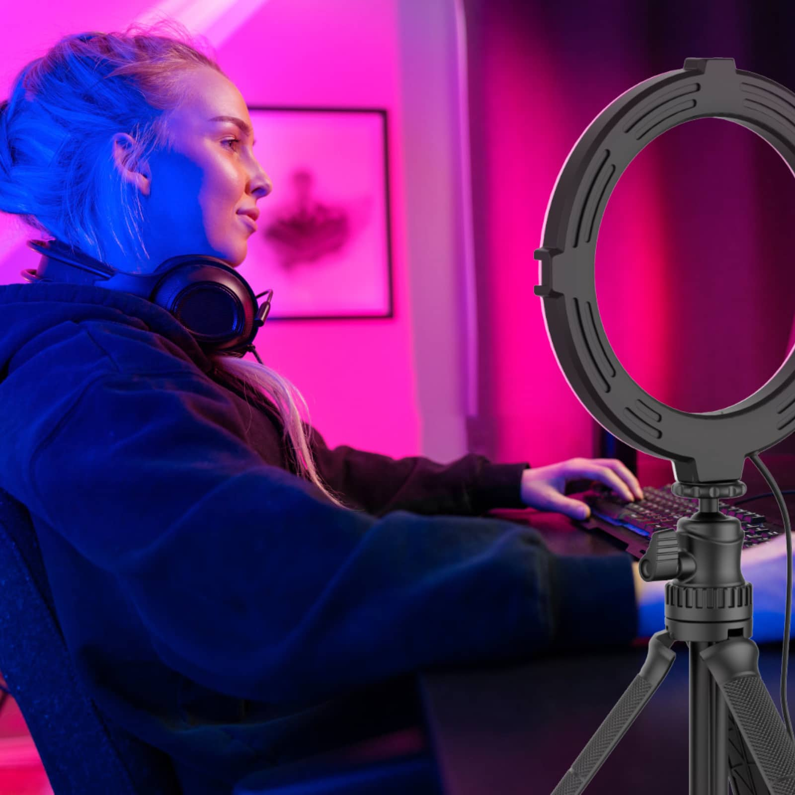 A woman with headphones sits in front of a computer with a ring light.