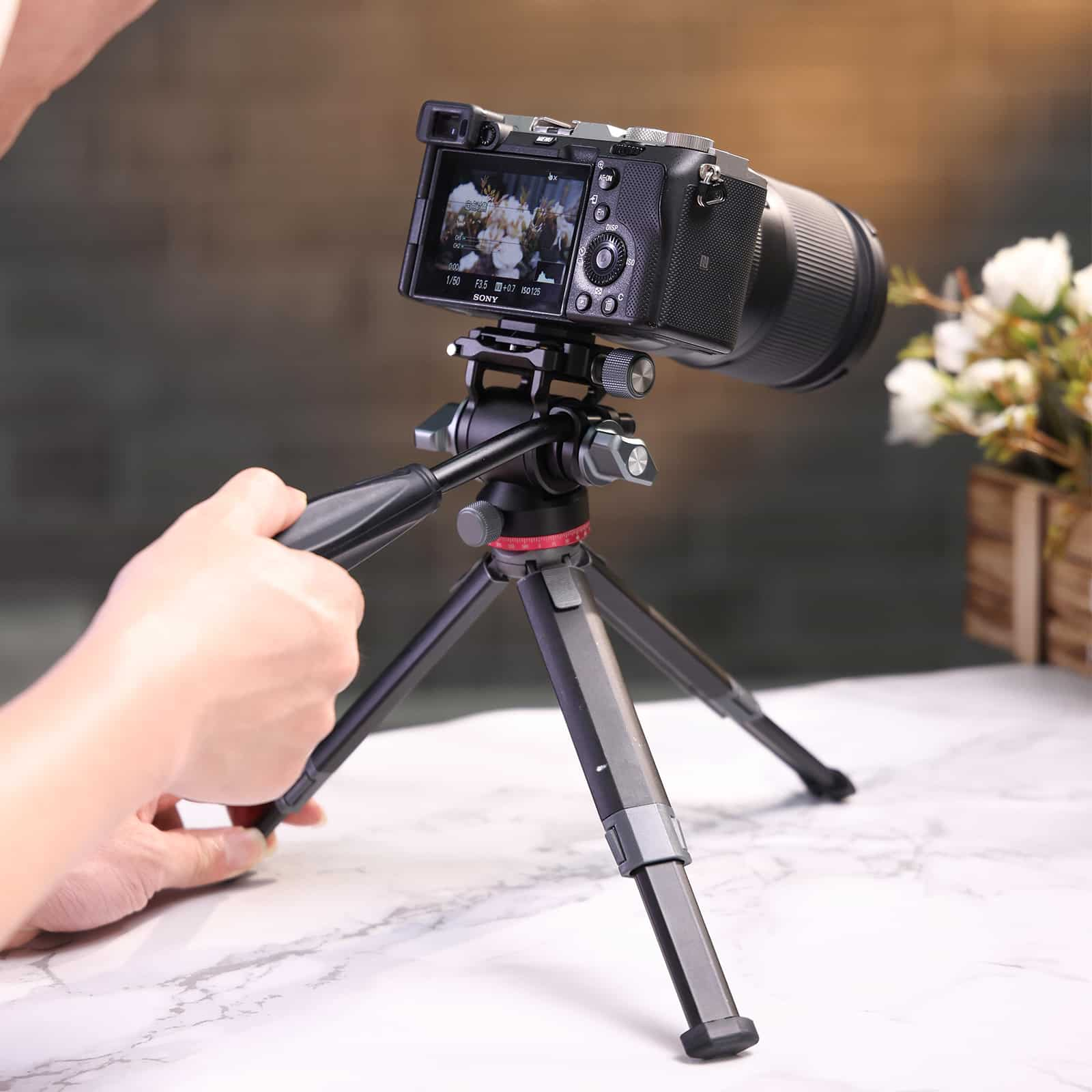 Aparat z obiektywem na statywie. Ręka trzyma statyw. Kwiaty w tle.