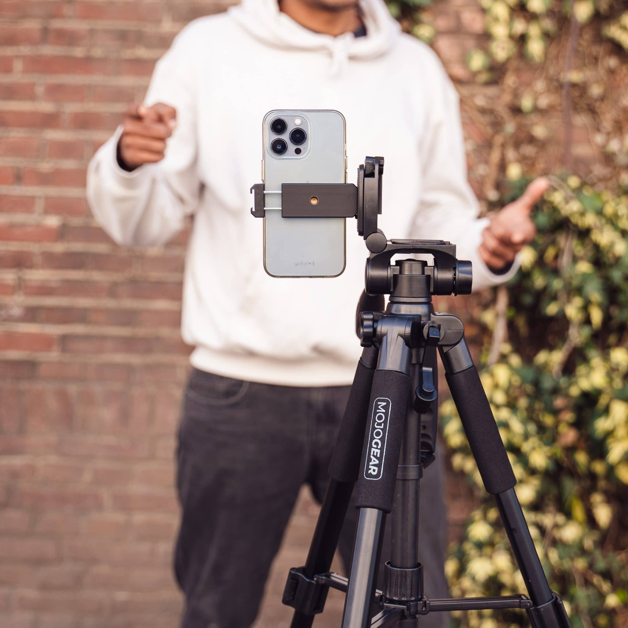 A person is filming with a phone mounted on a tripod. The background has a brick wall and foliage.
