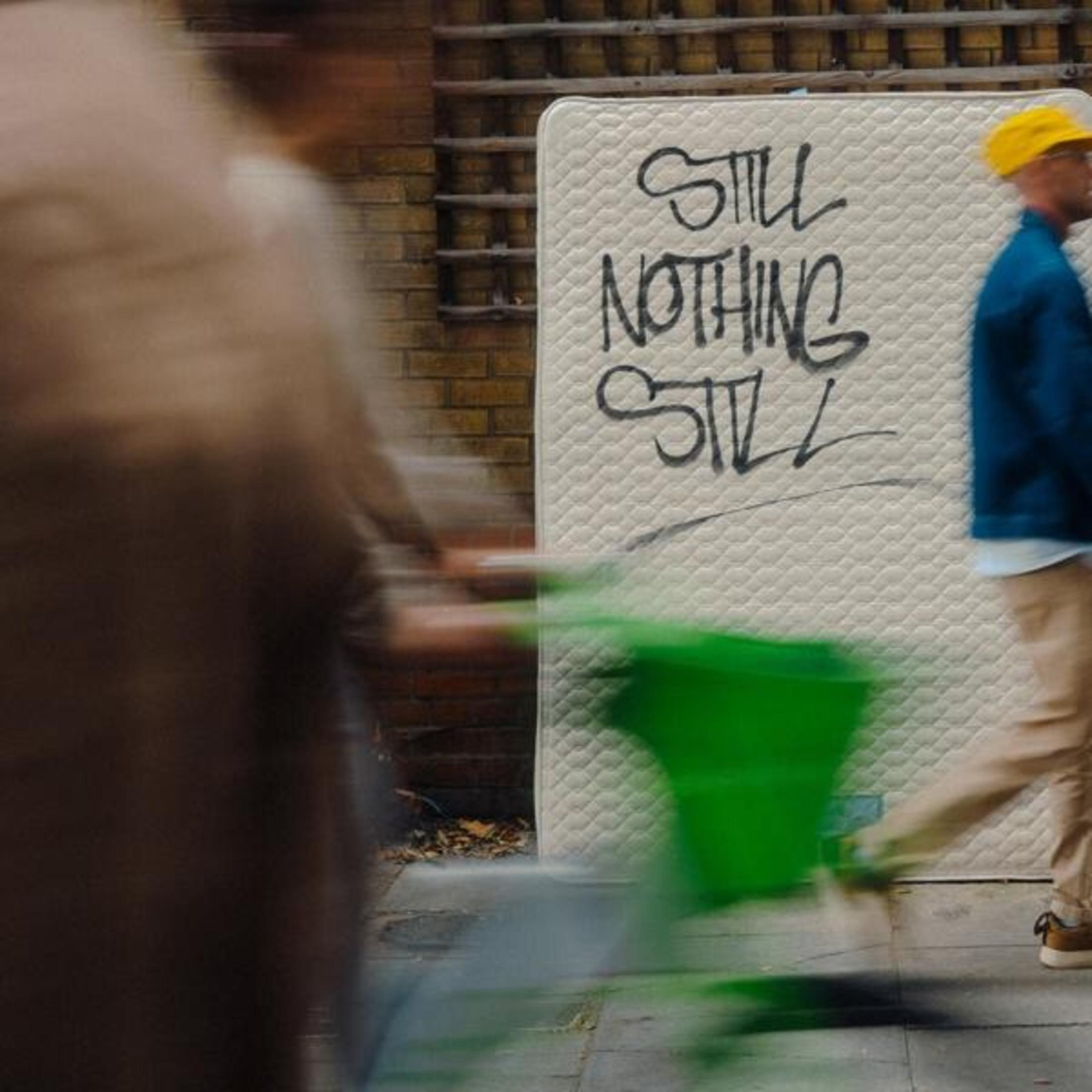 Une scène de rue avec un matelas et deux personnes qui passent. Les mots STILL NOTHING STILL sont écrits sur le matelas.
