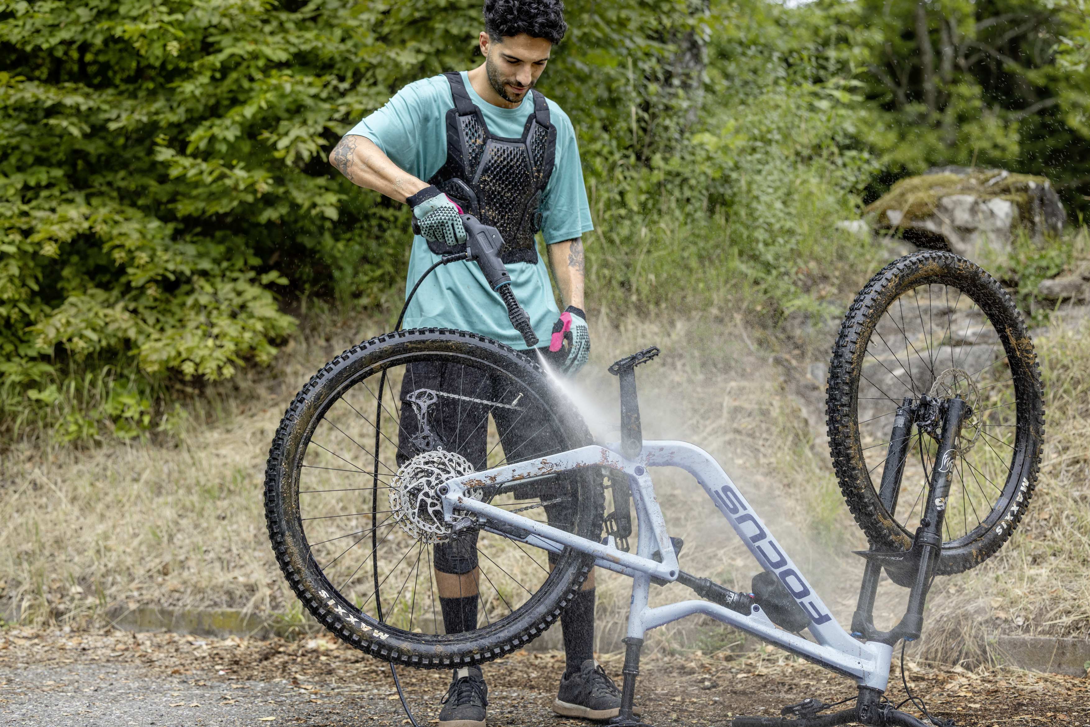 Man cleaning a blue mountain bike with a pressure washer. Green trees are in the background.