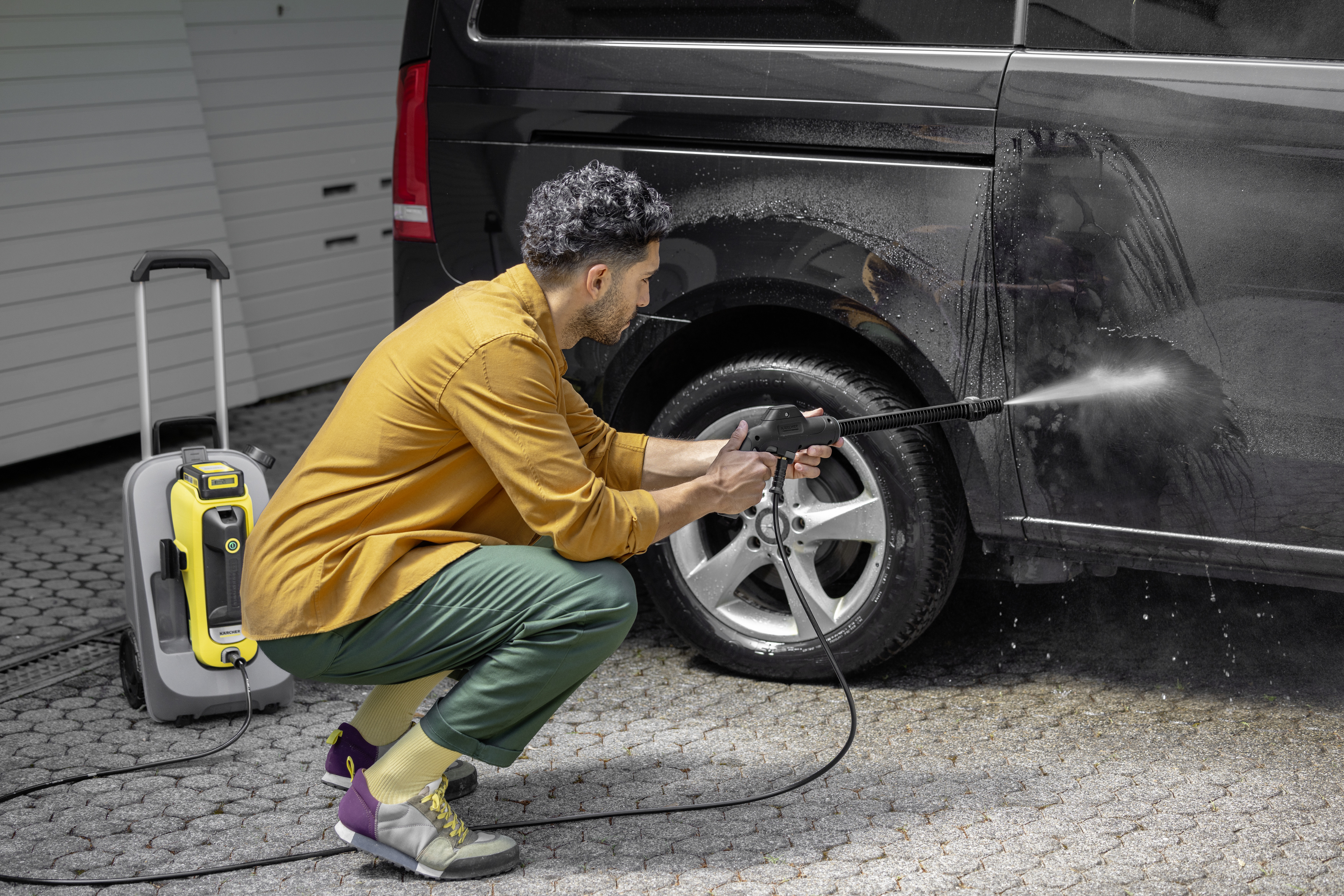 Mann wäscht einen schwarzen Transporter mit einem Hochdruckreiniger. Auto steht vor einer Garage.