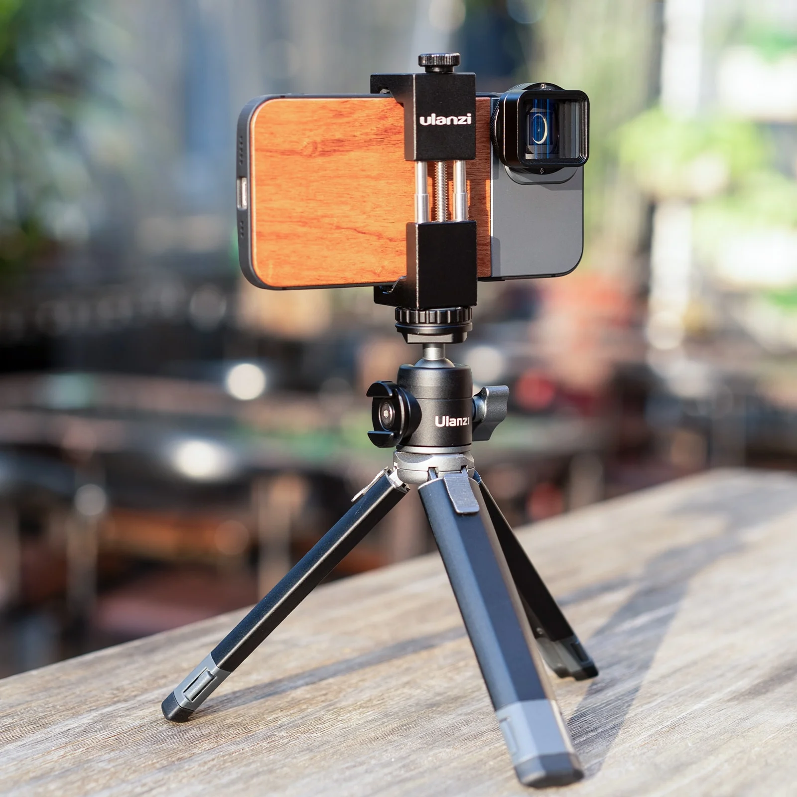 Un téléphone avec un objectif fixé à un trépied. Le trépied est sur une surface en bois, avec un arrière-plan flou d'un bâtiment.