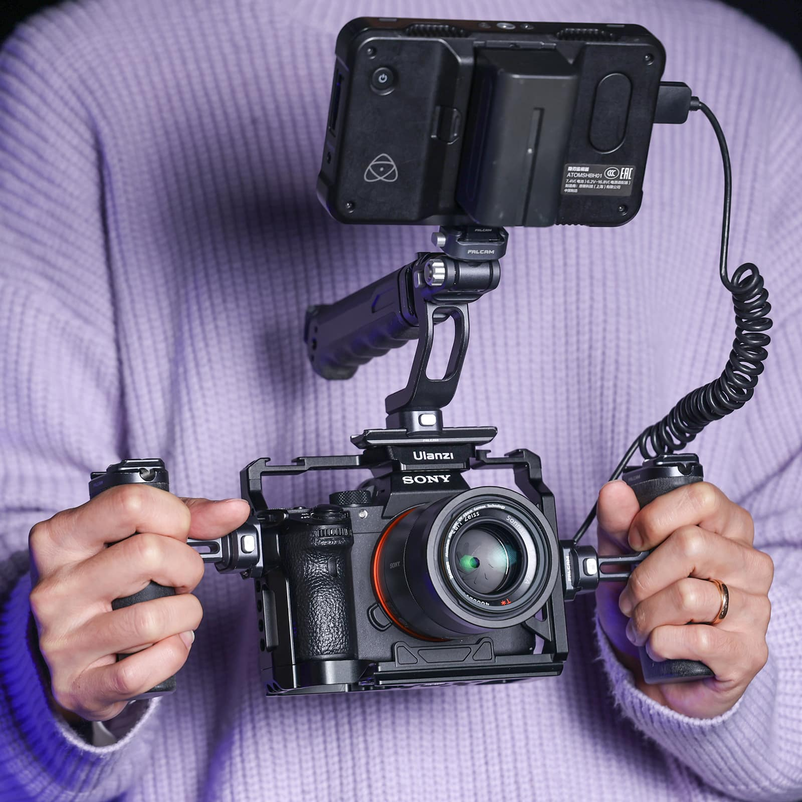 Persoon die een camerarig vasthoudt. De rig bevat een camera, monitor en handgrepen. Paarse achtergrond.