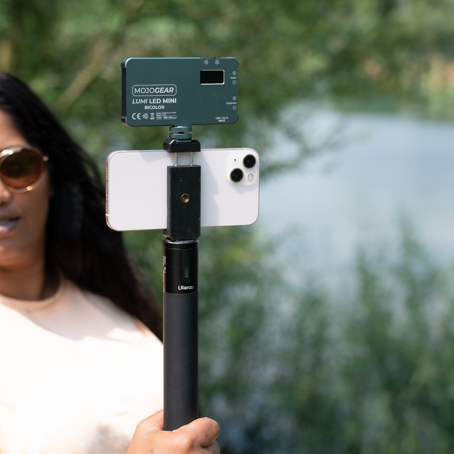 Een vrouw houdt een telefoon vast op een selfiestick met een lampje eraan. De achtergrond is een meer en groen.